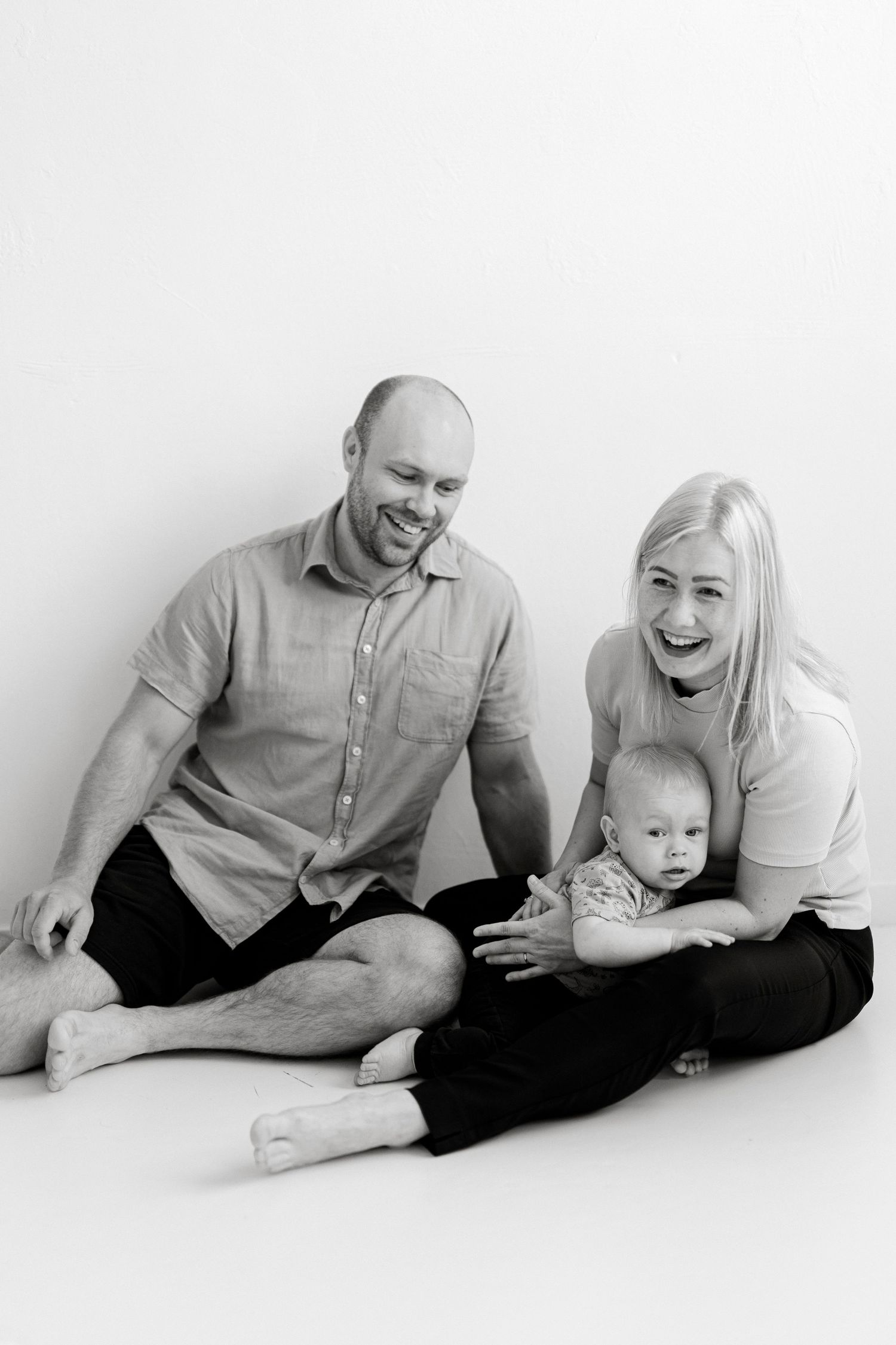 A joyful family portrait in black and white showing parents and baby sitting together on a white background.