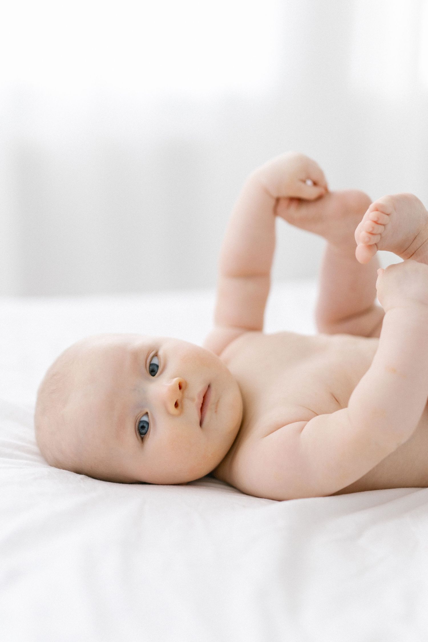 A cute naked baby with blue eyes lying on a white bed, holding their feet up in the air.