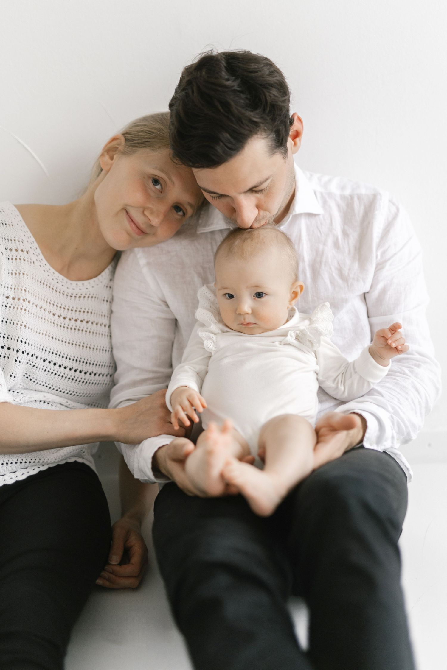 Young family with mother, father, and baby dressed in white sitting together against a white wall.