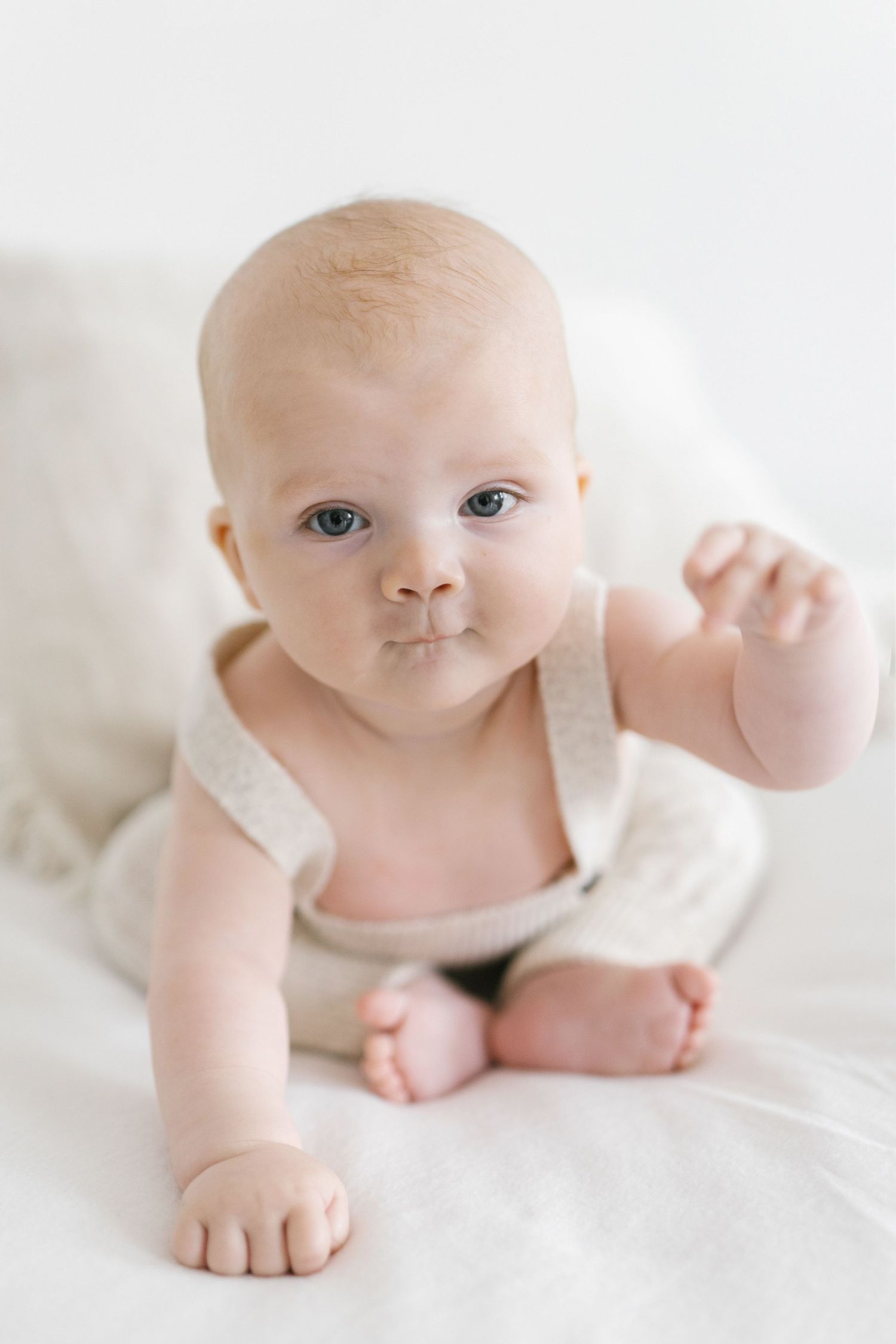 Adorable baby in a neutral knit romper doing tummy time on a white blanket, looking curiously at the camera.