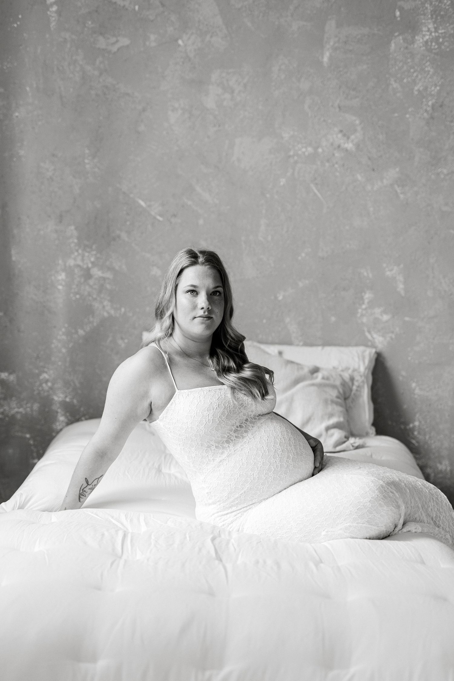 Black and white maternity photo of a pregnant woman in white lace dress sitting on a bed with pillows.