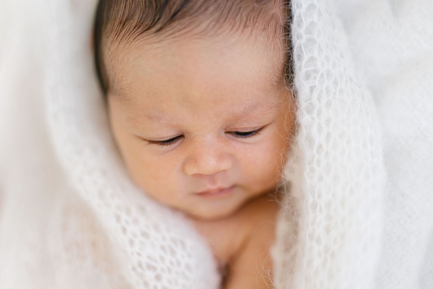 Peaceful newborn baby wrapped in a soft white knit blanket, eyes closed, with delicate features in a close-up portrait.