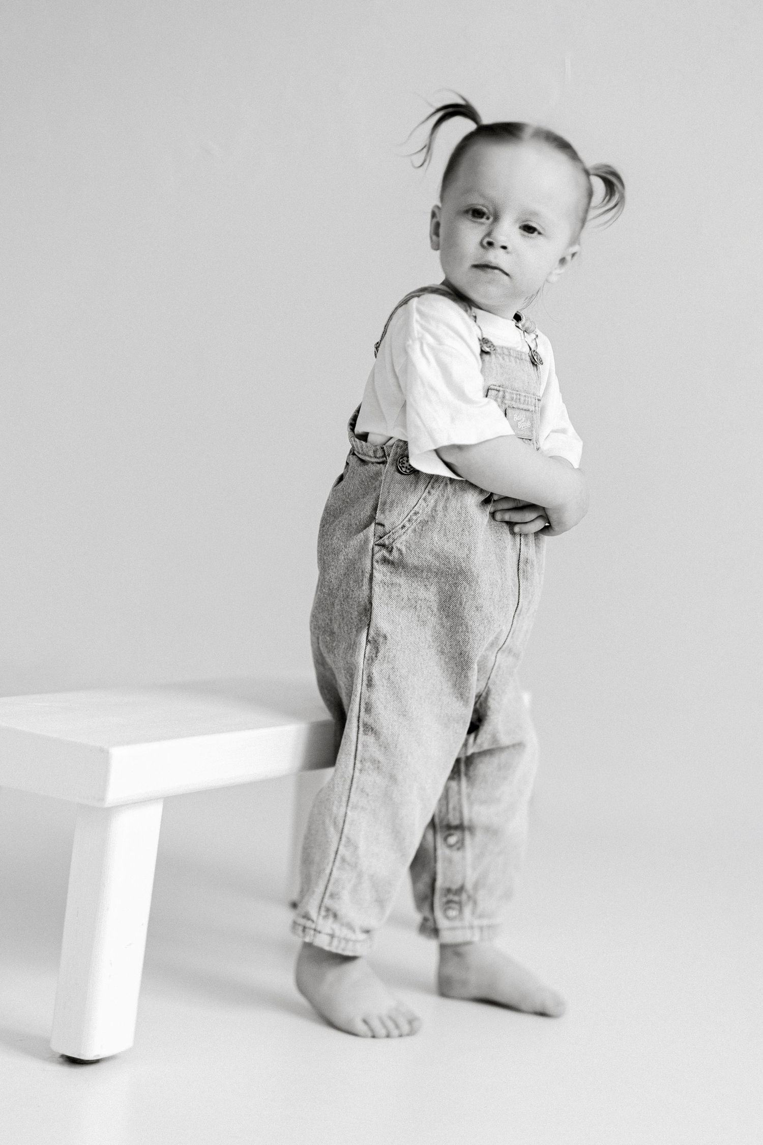 Black and white photo of a cute toddler in denim overalls with pigtails, standing barefoot with arms crossed.