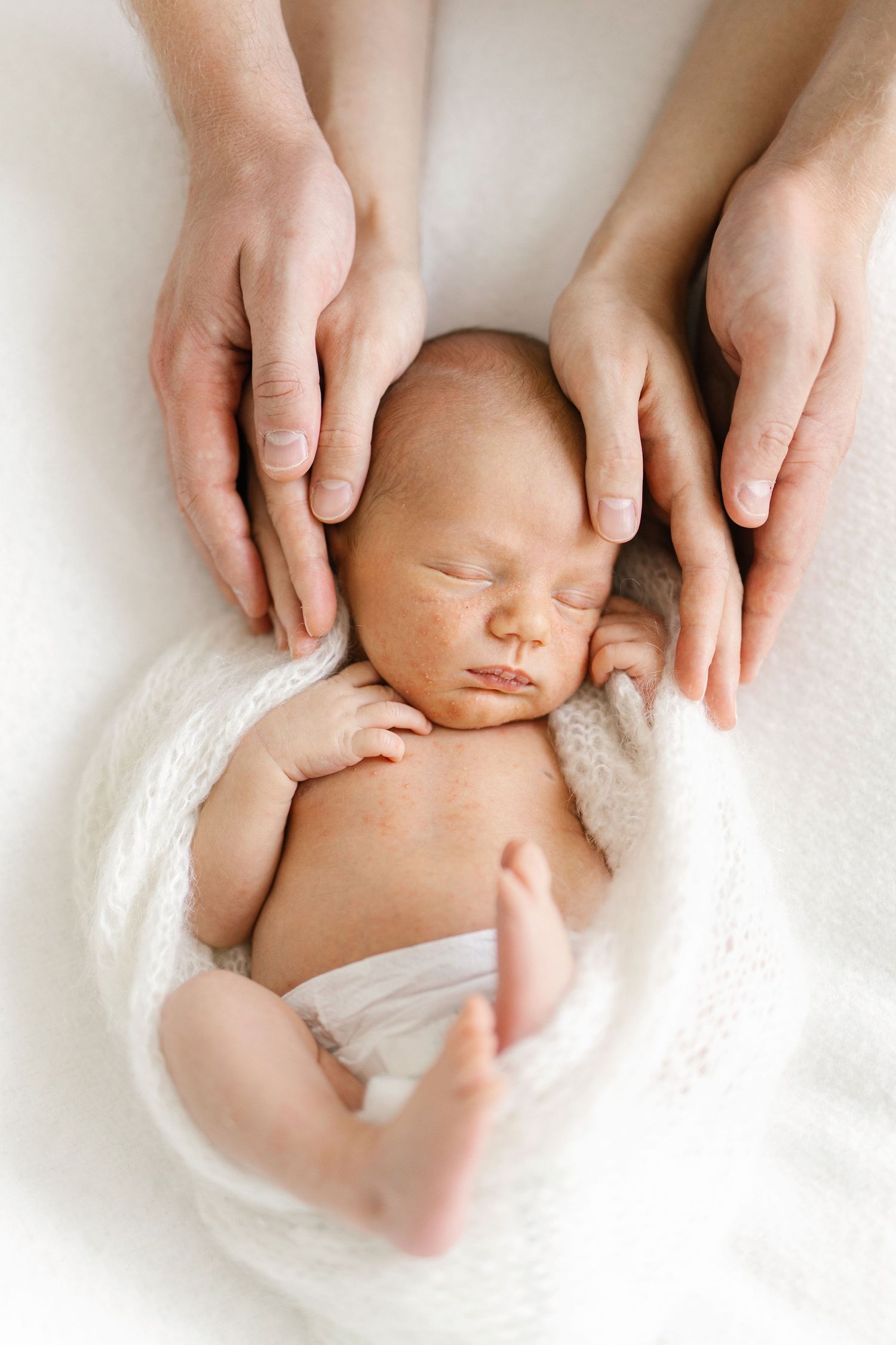 Newborn baby sleeping peacefully wrapped in white, cradled by parents hands on soft white background.