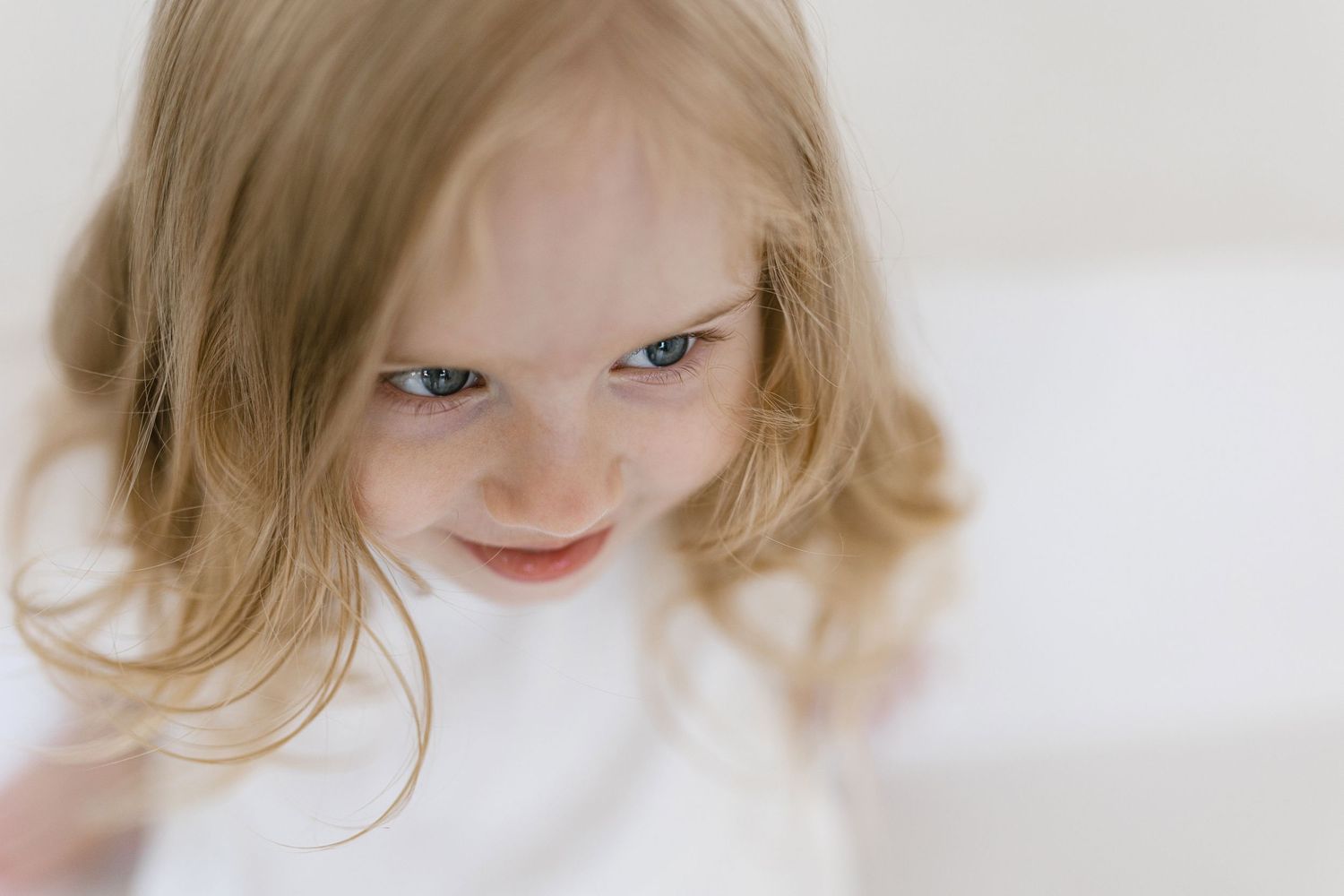 Blonde toddler girl with blue eyes giving a mischievous smirk, looking down at camera against white background.
