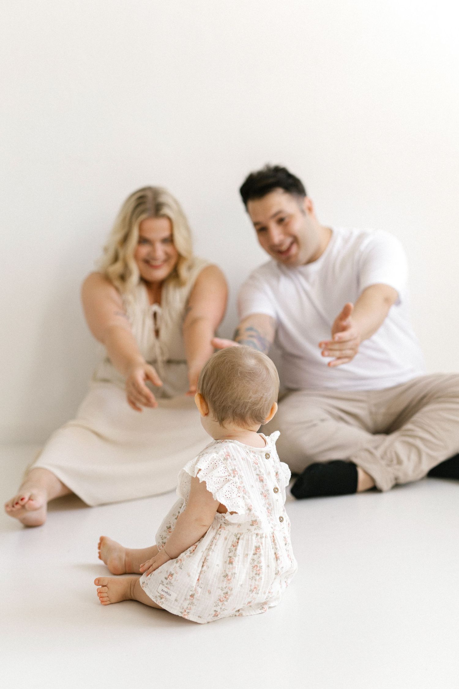 A happy couple sits on a white floor reaching toward their baby girl in a floral dress during a family photo session.