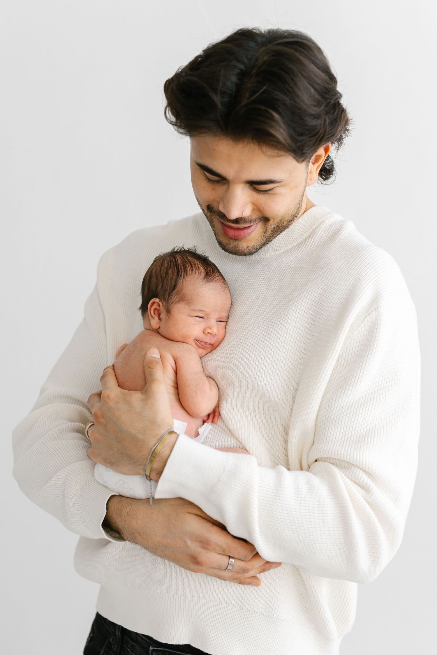 Smiling young father in white sweater tenderly holds his newborn baby against a white background.