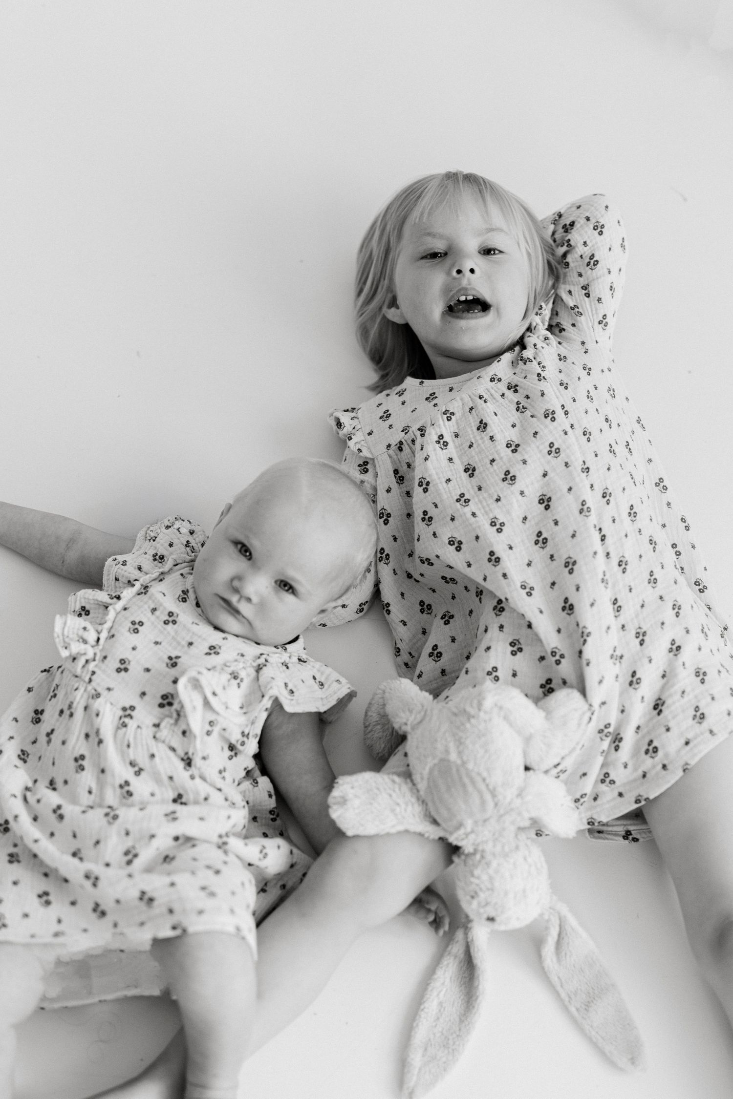 Two young sisters in matching floral dresses pose together, with a baby holding a stuffed bunny toy.