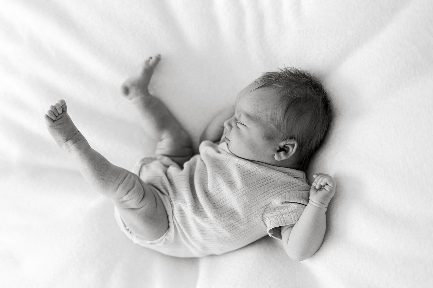 Black and white photo of a sleeping newborn baby lying on a soft white blanket with arms stretched out.