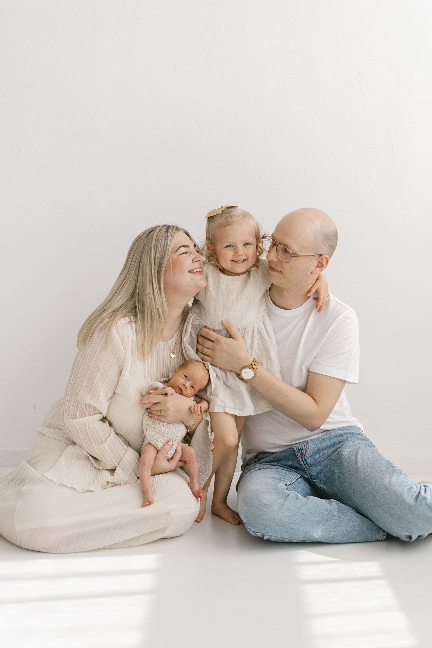 Happy family with newborn baby and toddler daughter posing together on white studio floor.