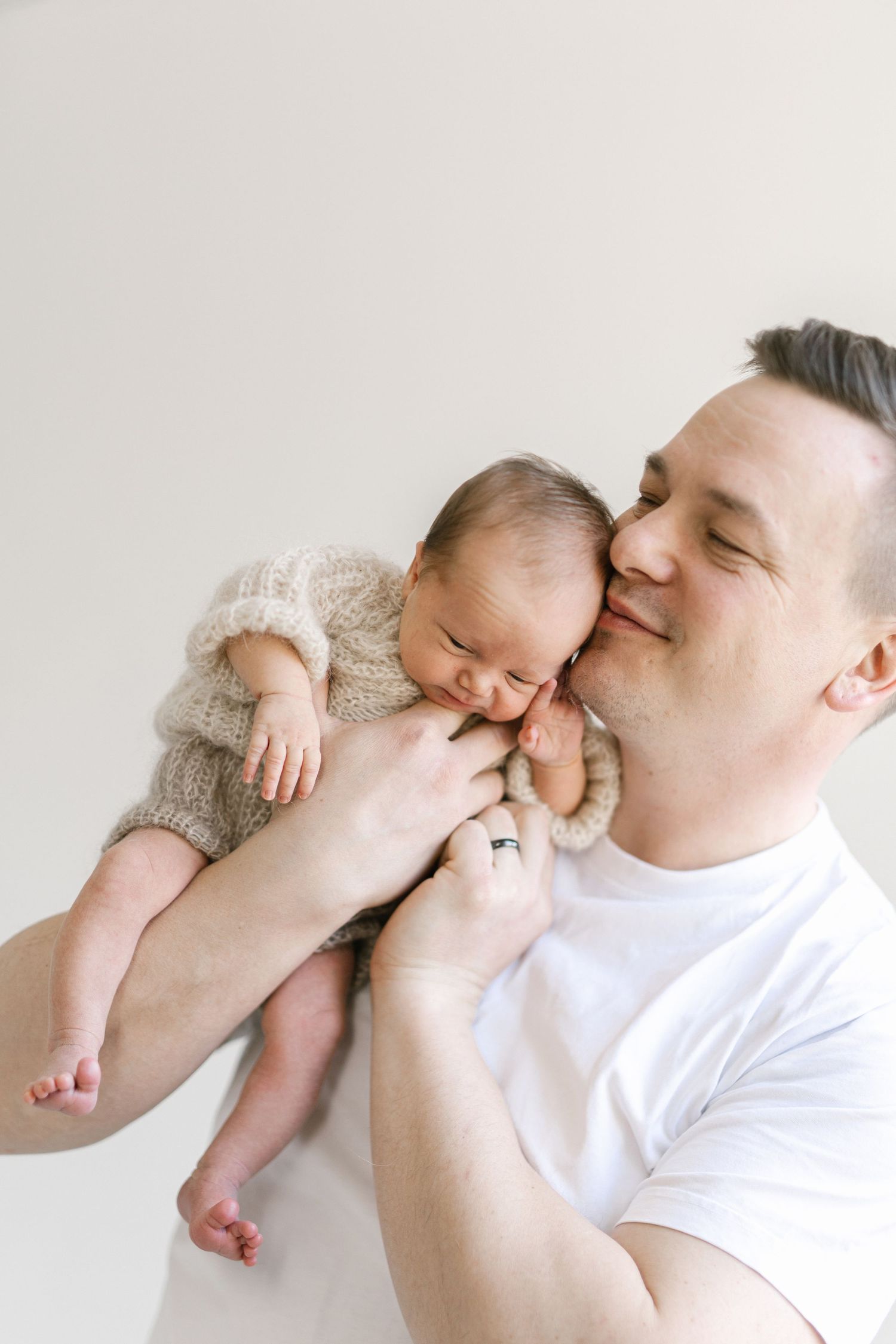 A father tenderly kisses his sleeping newborn baby dressed in a knit romper against a light background.
