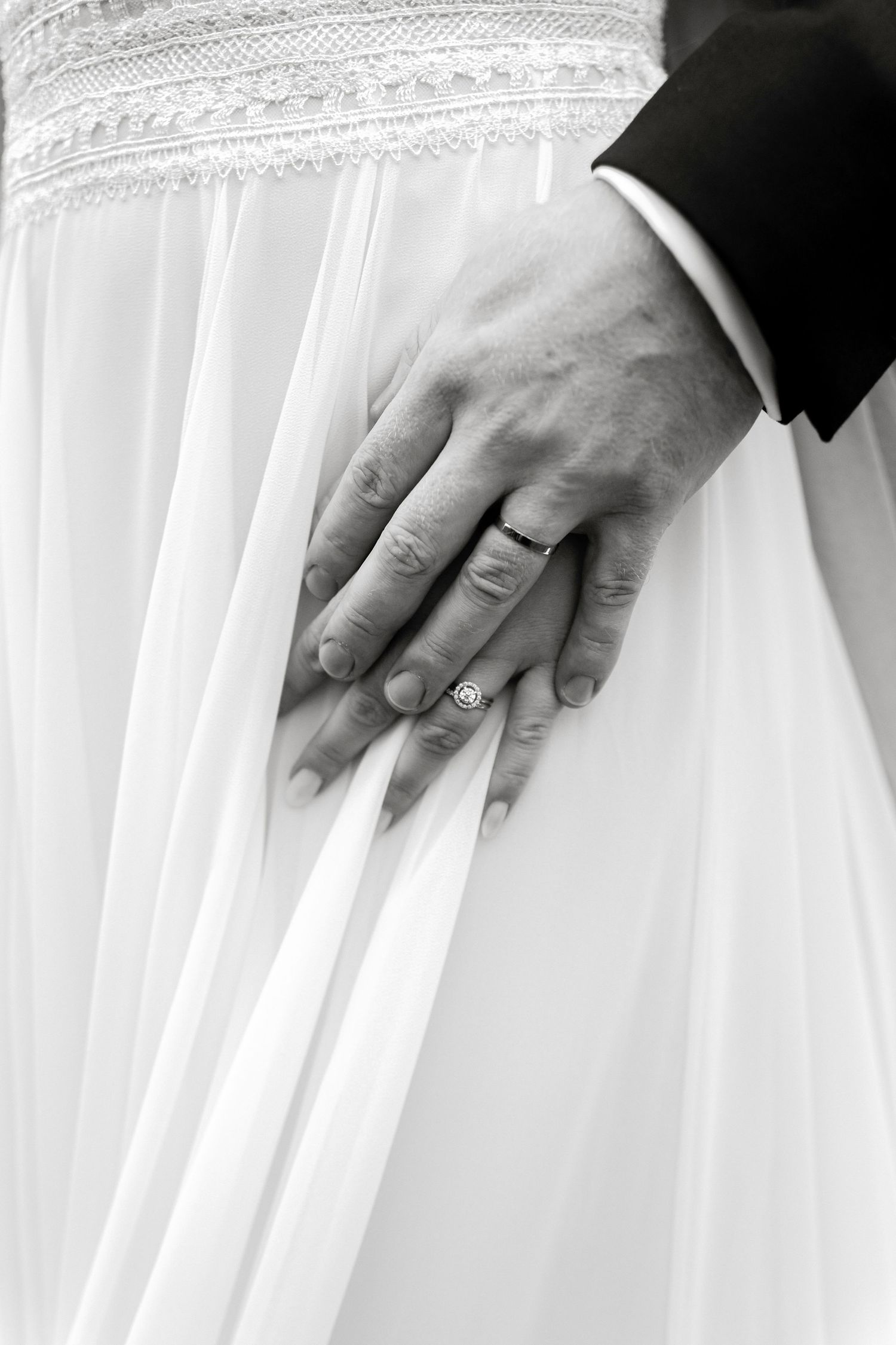Black and white photo of newlyweds hands with wedding rings resting on white bridal gown.