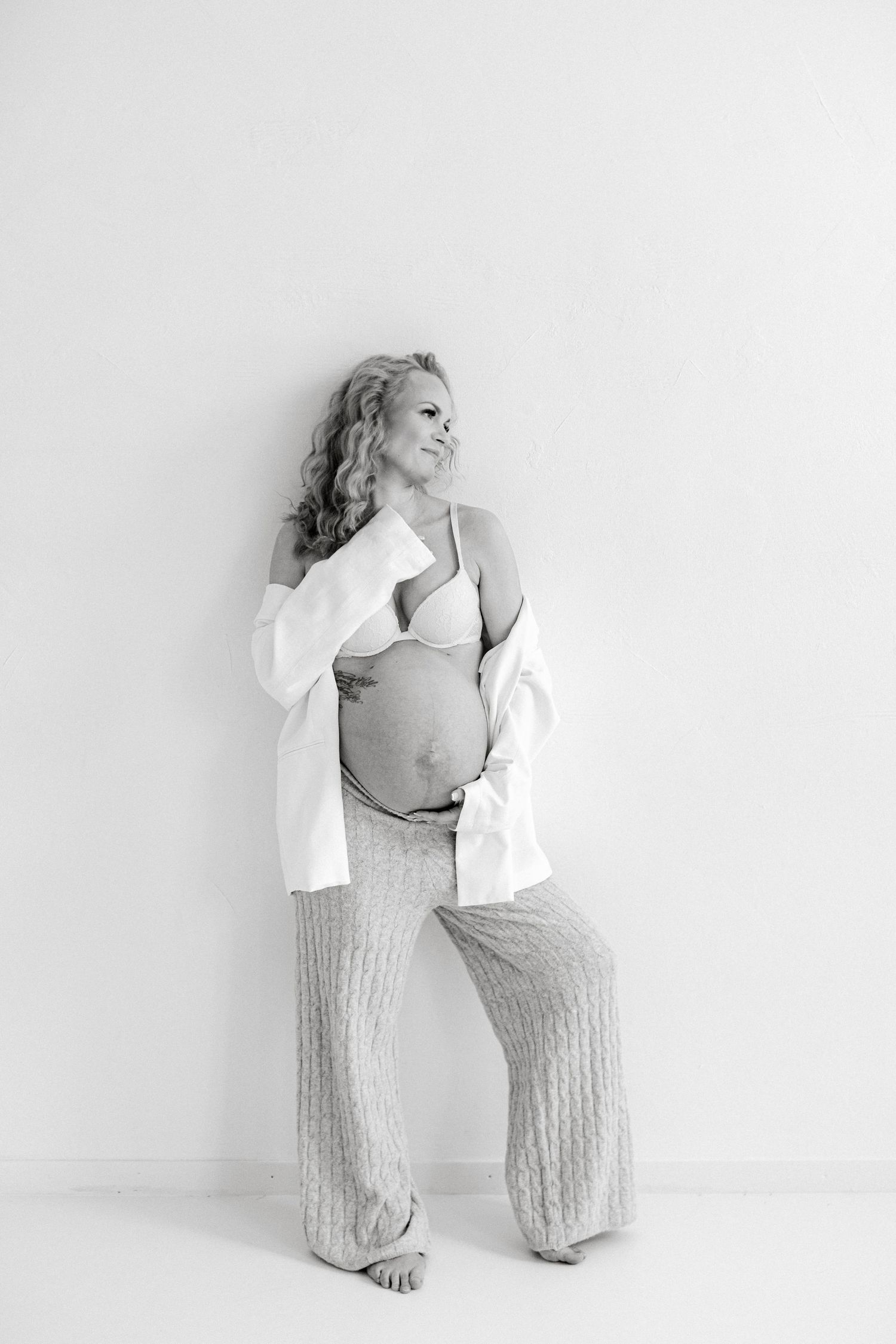 Black and white maternity portrait of a pregnant woman with curly hair leaning on a wall, wearing an open white shirt and knit pants.