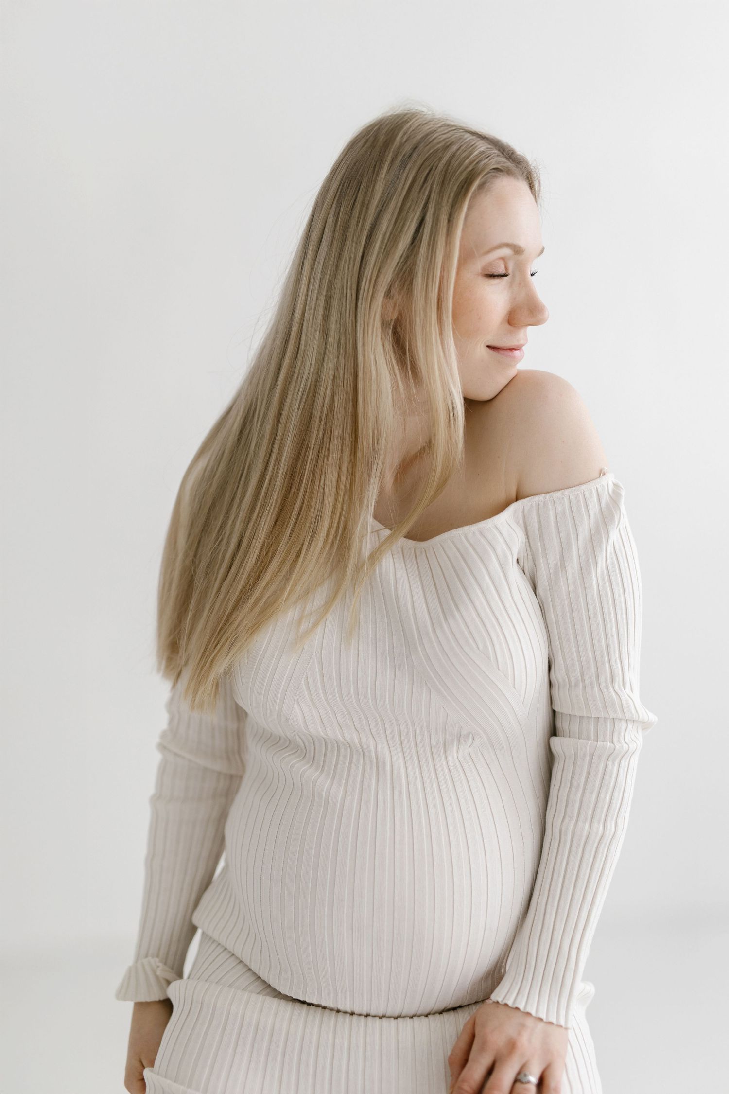 Pregnant woman in cream ribbed off-shoulder top smiling downward, showcasing baby bump against white background.