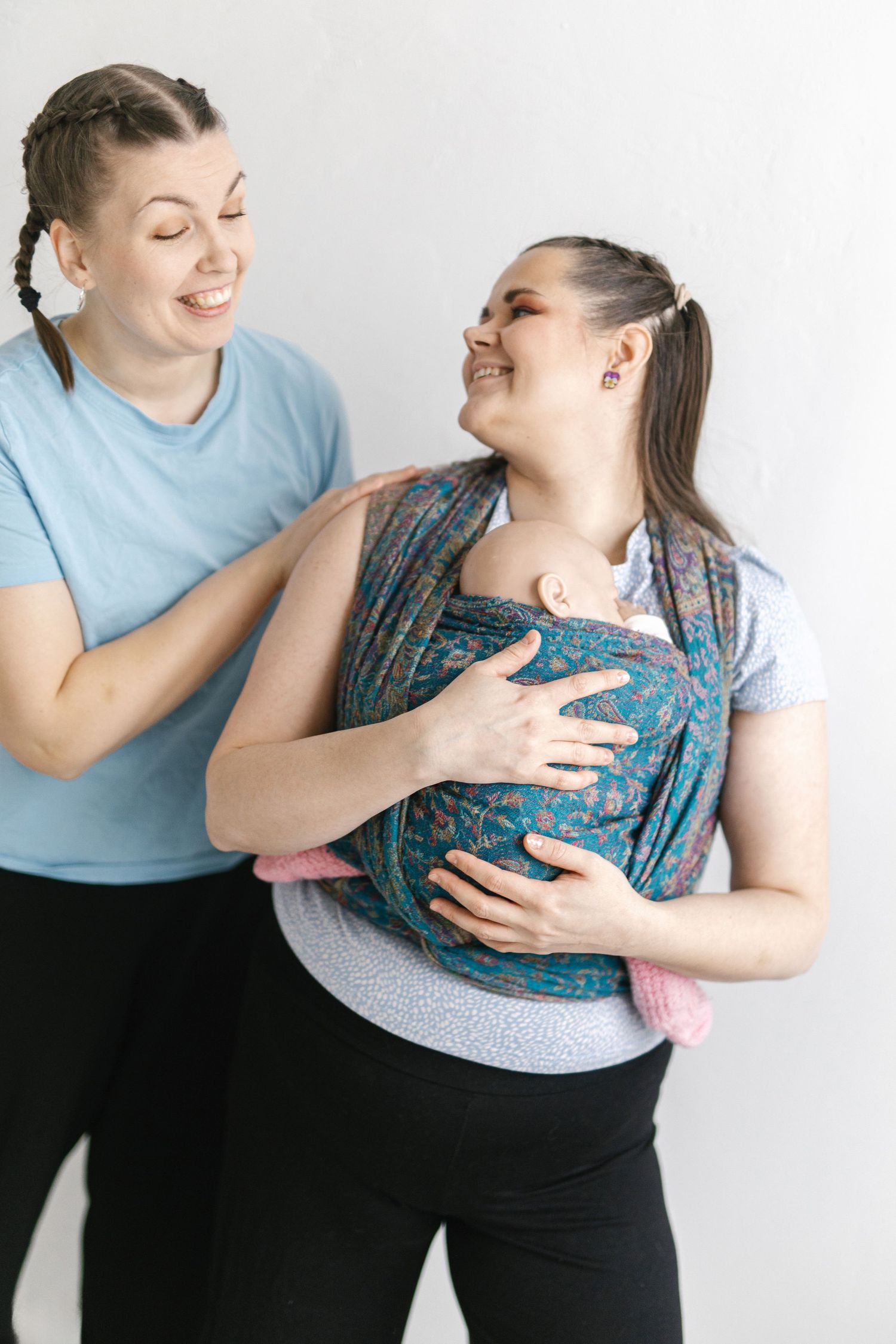 Two women smiling, one wearing a floral baby carrier with a newborn, while the other assists her.