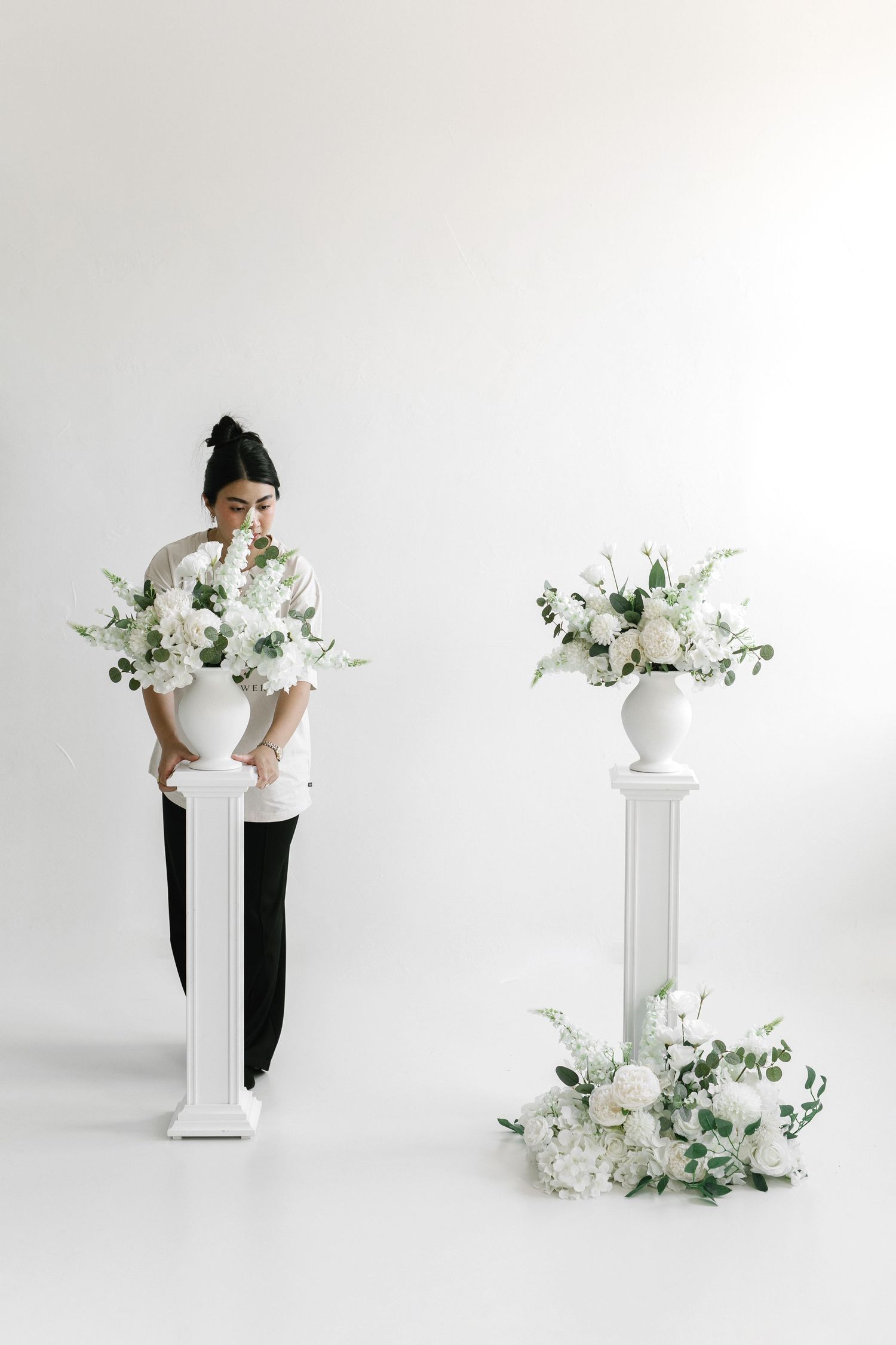 Woman in black and white outfit arranging white floral arrangements on decorative pedestals against white background.