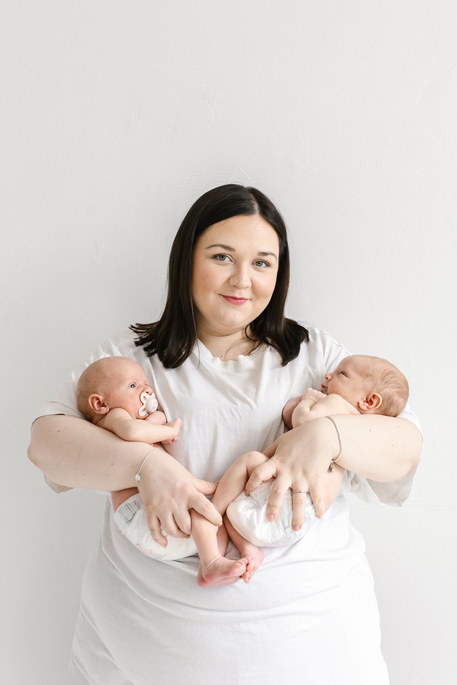 A mother in a white dress holds two newborn twins, one in each arm, against a white background.