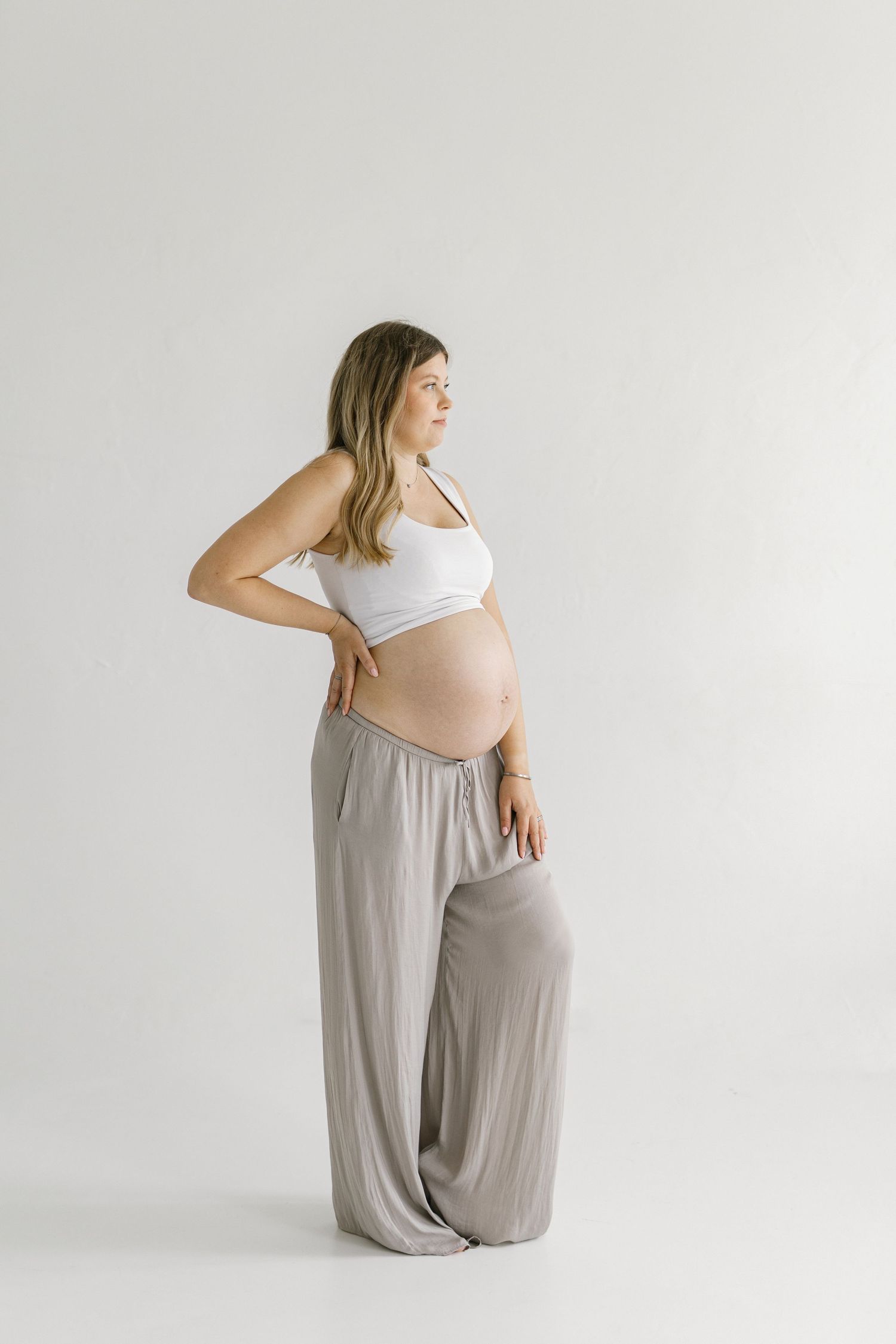 Pregnant woman in white crop top and taupe wide-leg pants posing sideways, showing baby bump against white background.