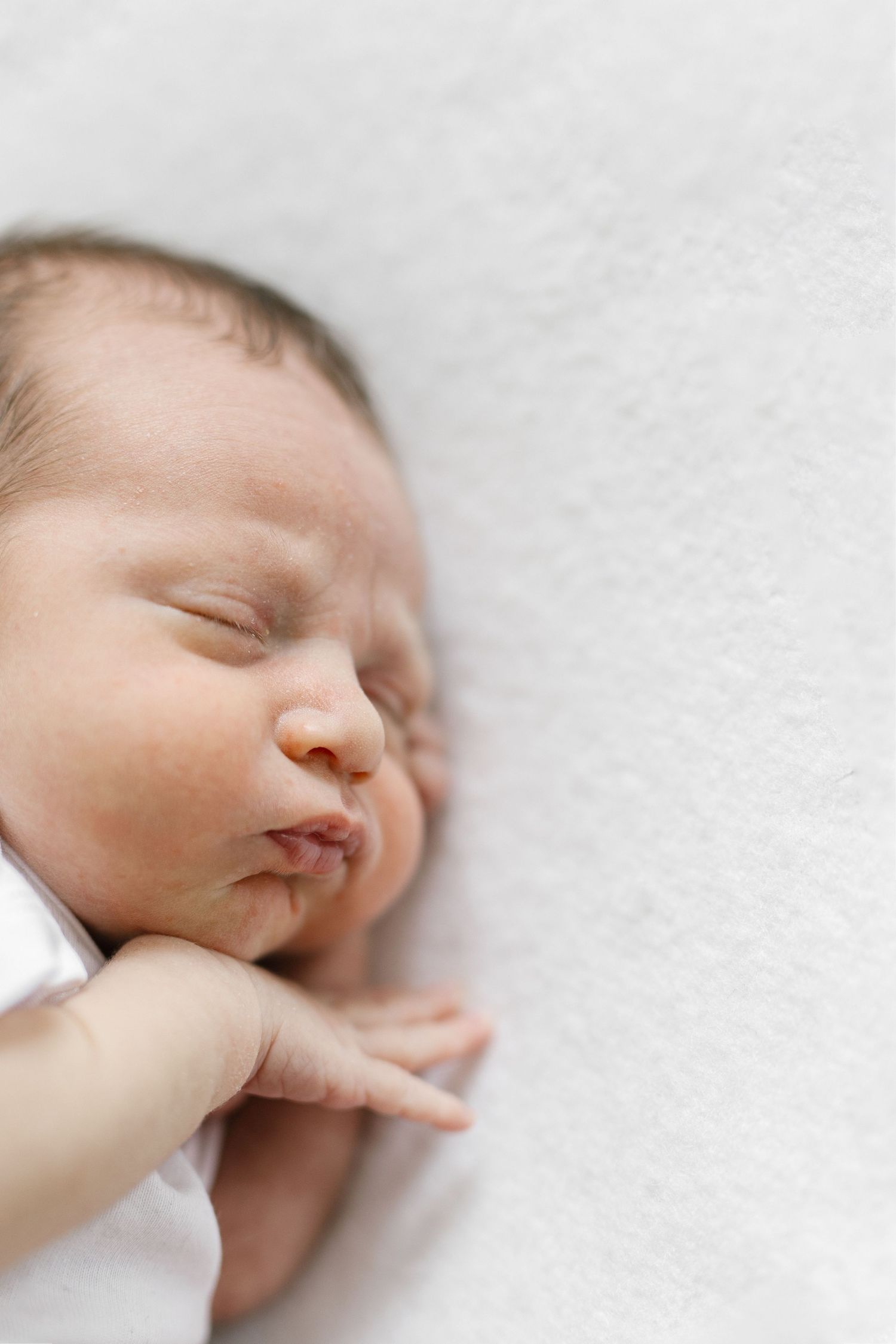 Peaceful newborn baby sleeping on white blanket, eyes closed, tiny hand visible, wearing white onesie.
