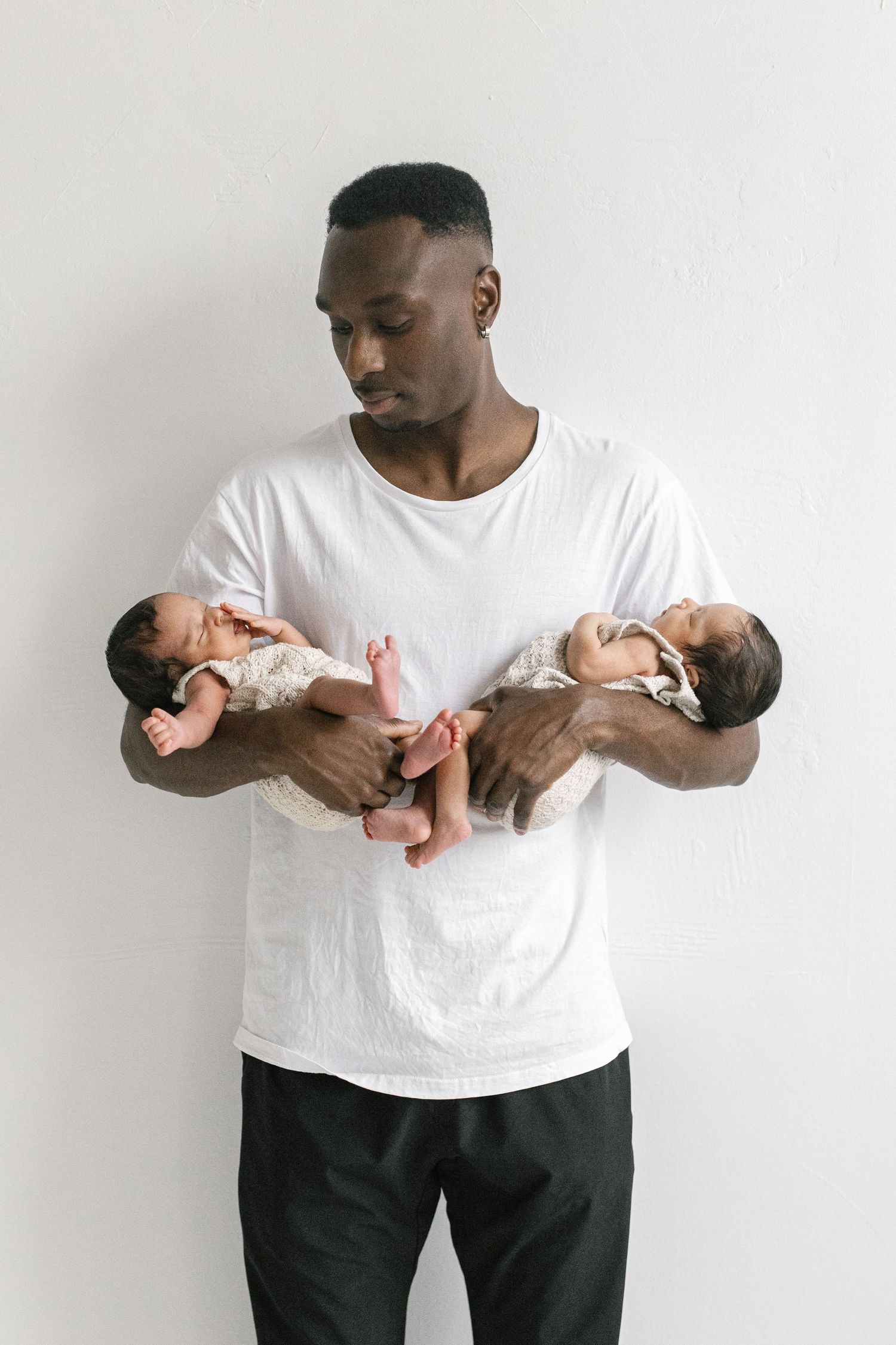 A proud Black father in a white t-shirt holds two sleeping newborn twins in his arms against a white wall.