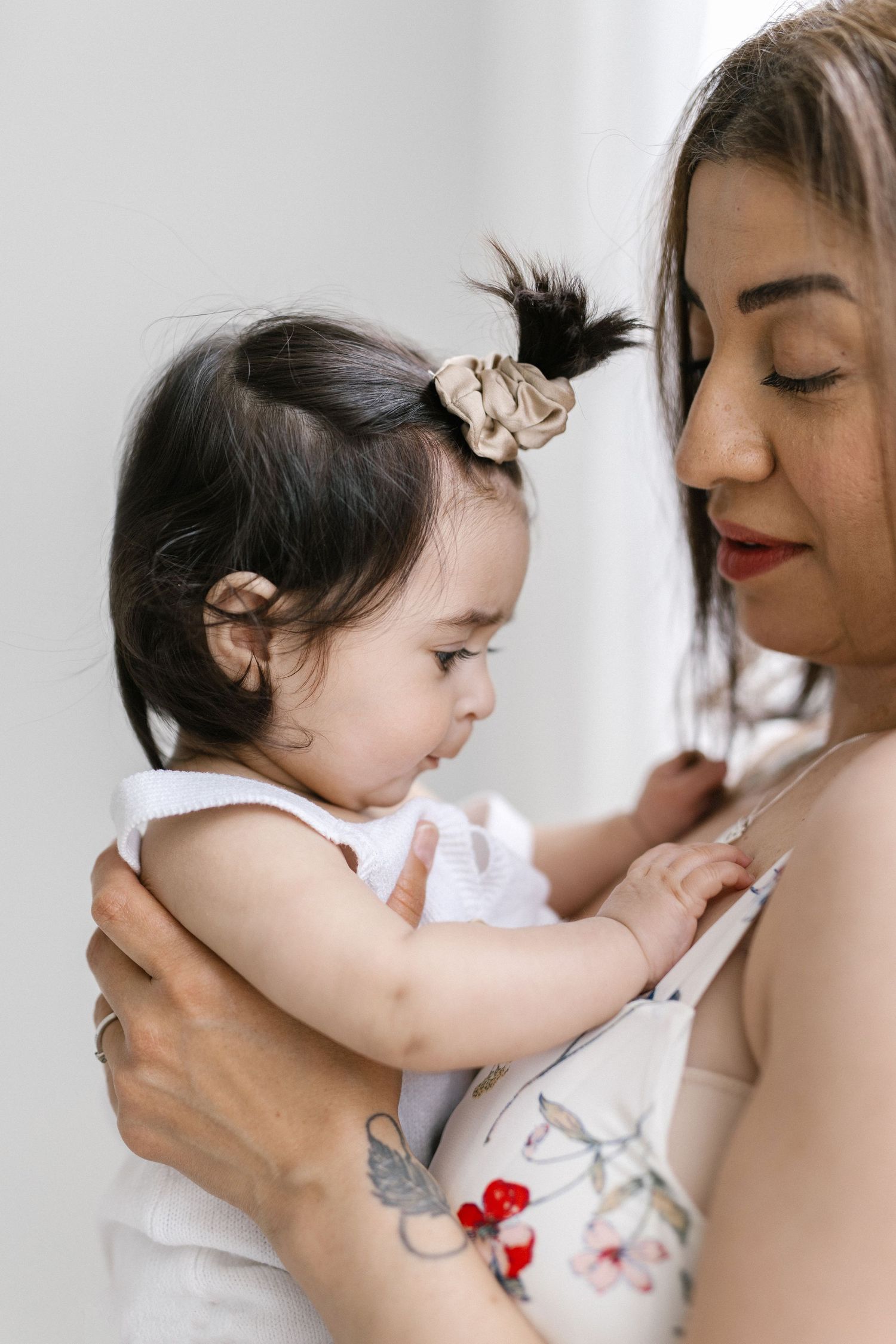 A mother with a floral tattoo tenderly holds her baby girl with a scrunchie and hearing aid in her hair.