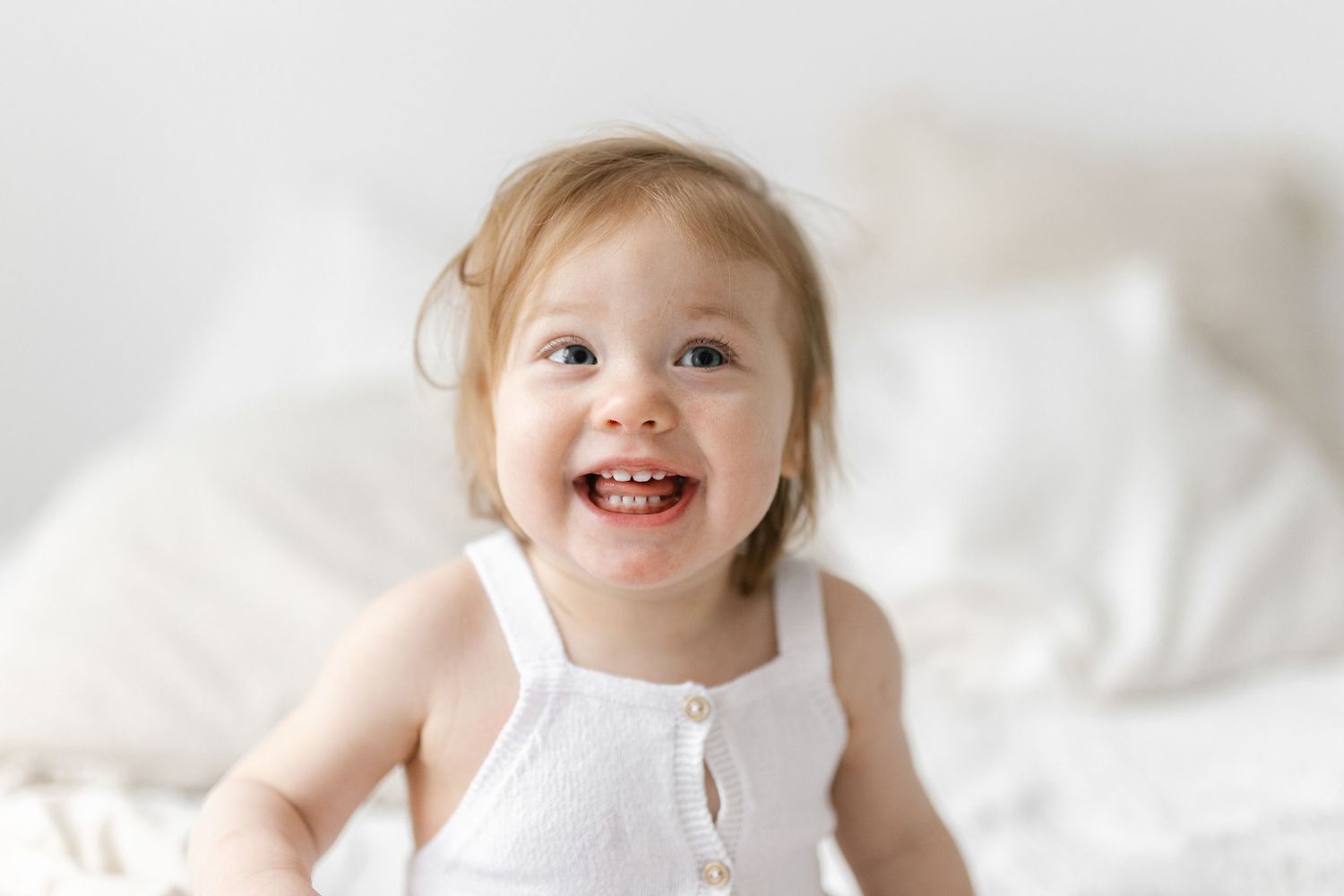 Smiling toddler girl in white dress sitting on white bed, happy baby with rosy cheeks in bright airy room.