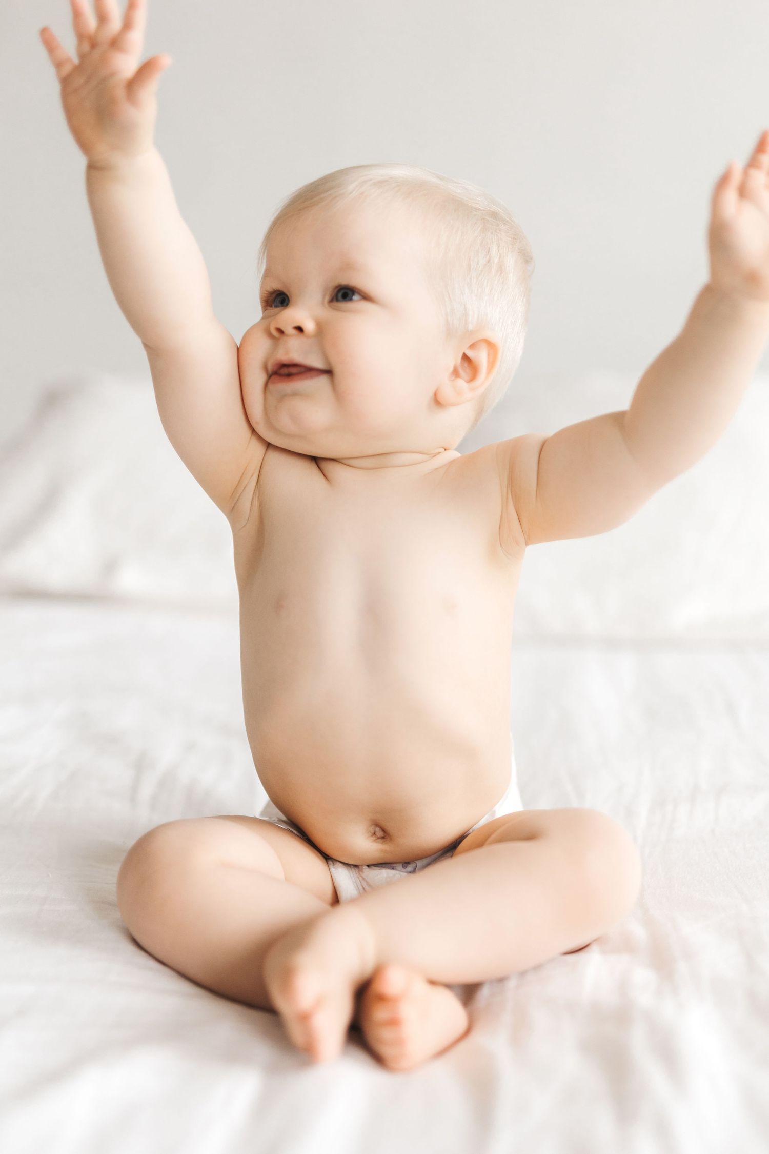 Happy baby in diaper sitting cross-legged on white bed with arms raised, smiling and looking to the side.