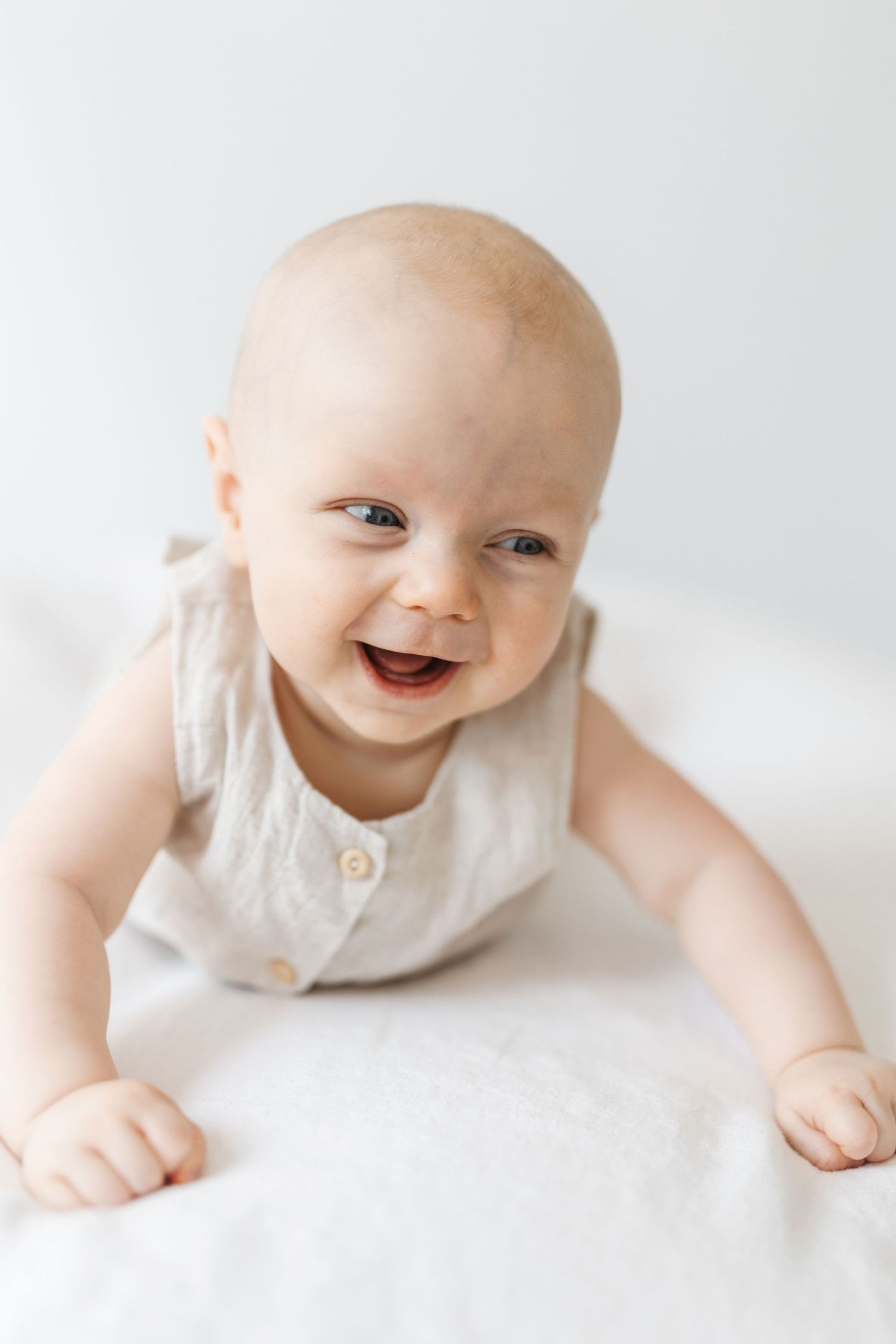 A happy baby in a beige linen romper doing tummy time on a white surface, smiling brightly at the camera.