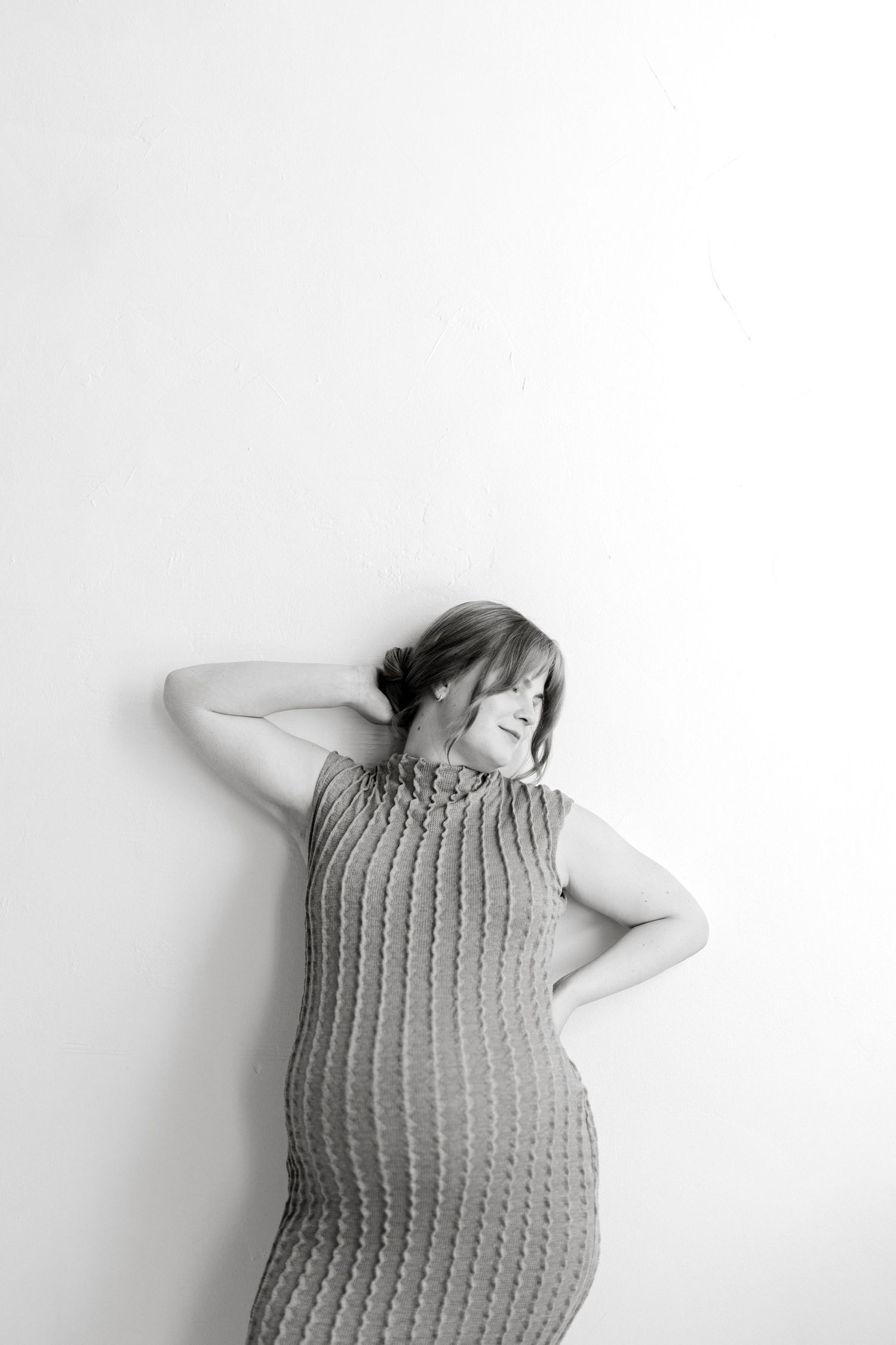 Black and white portrait of a pregnant woman in a fitted ribbed knit dress leaning against a white wall with one arm behind her head.