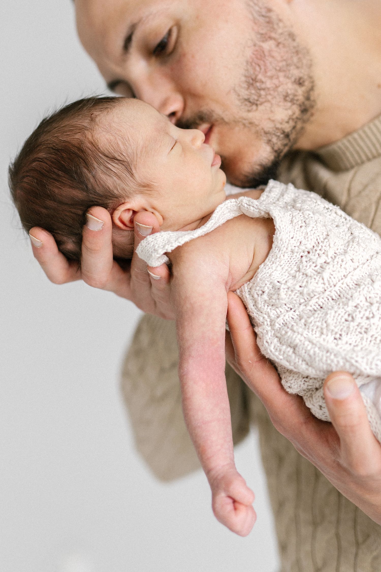 A father tenderly kisses his newborn baby dressed in a white knit outfit while cradling them in his hands.