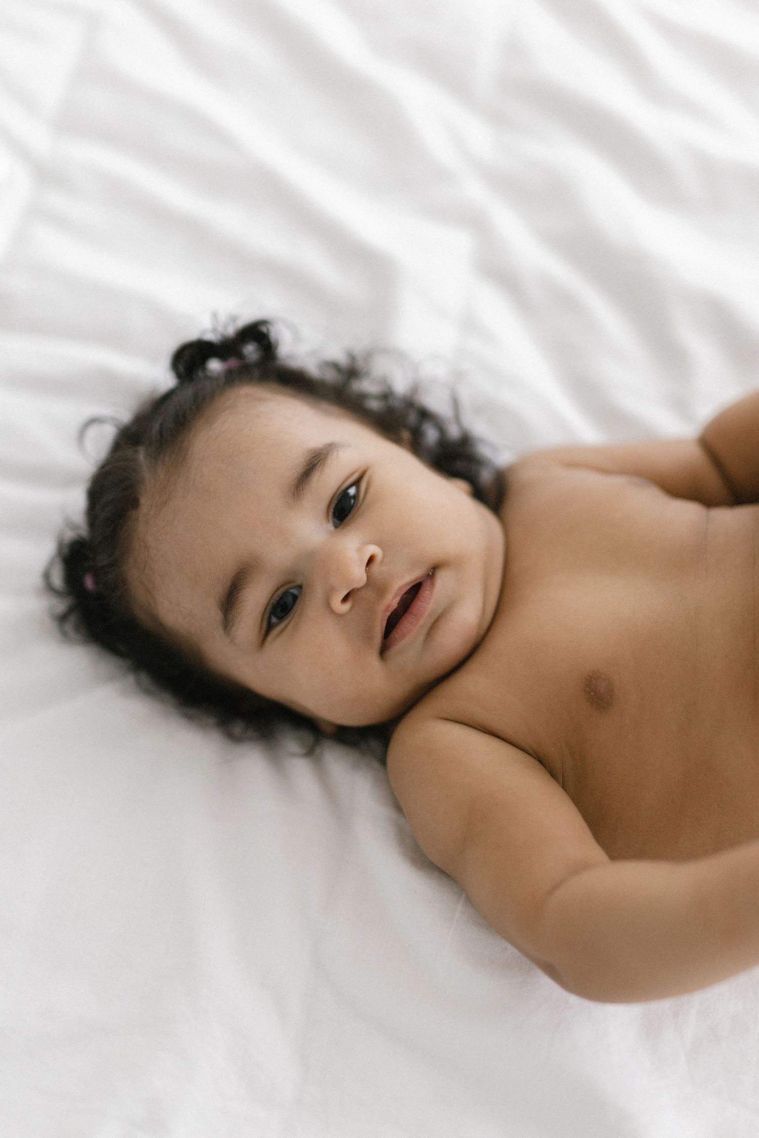 Adorable baby with curly hair lying on white bedding, looking up with curious eyes.