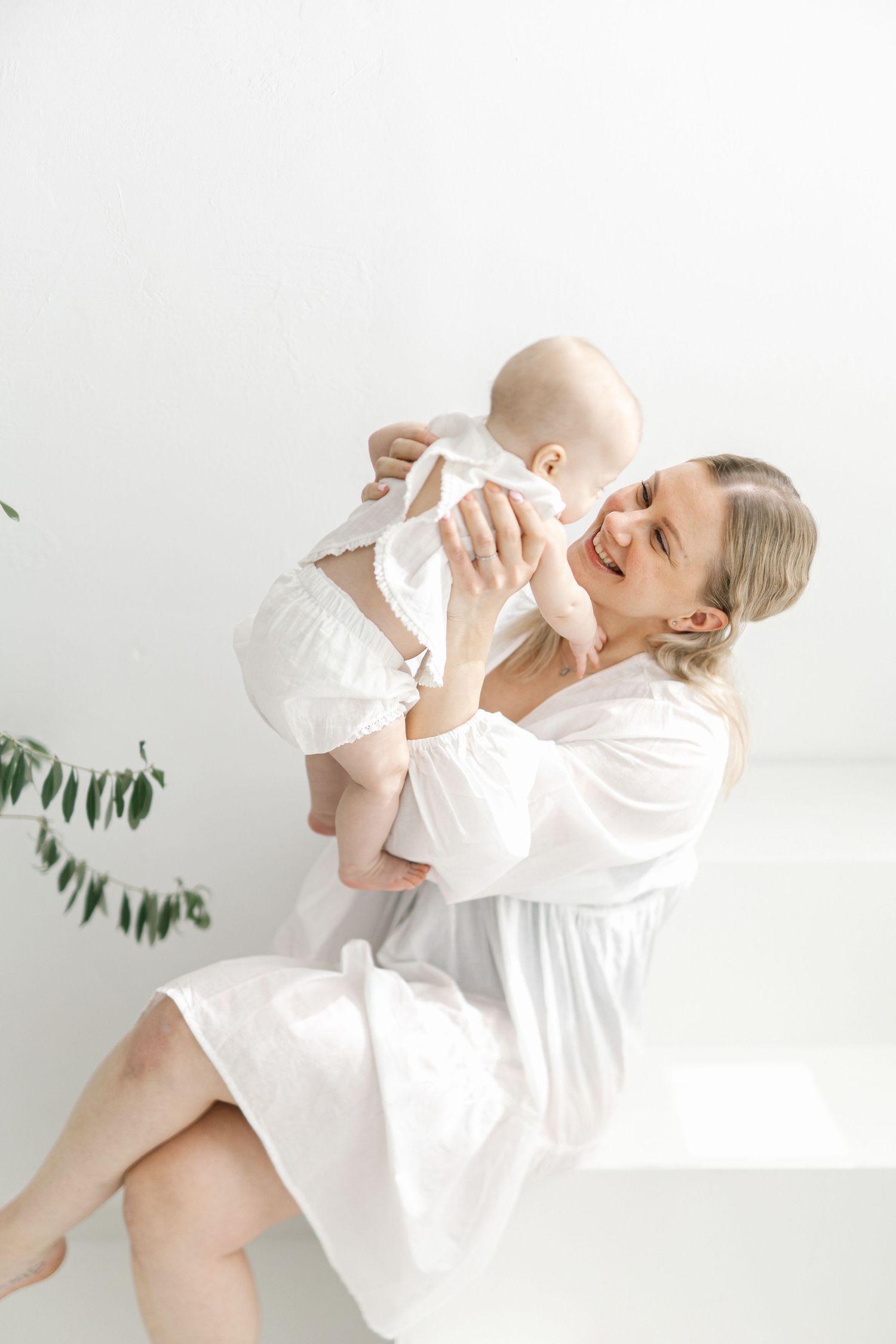 Happy mother in white dress holding and smiling at her baby, sharing a joyful moment in a bright, minimalist room.