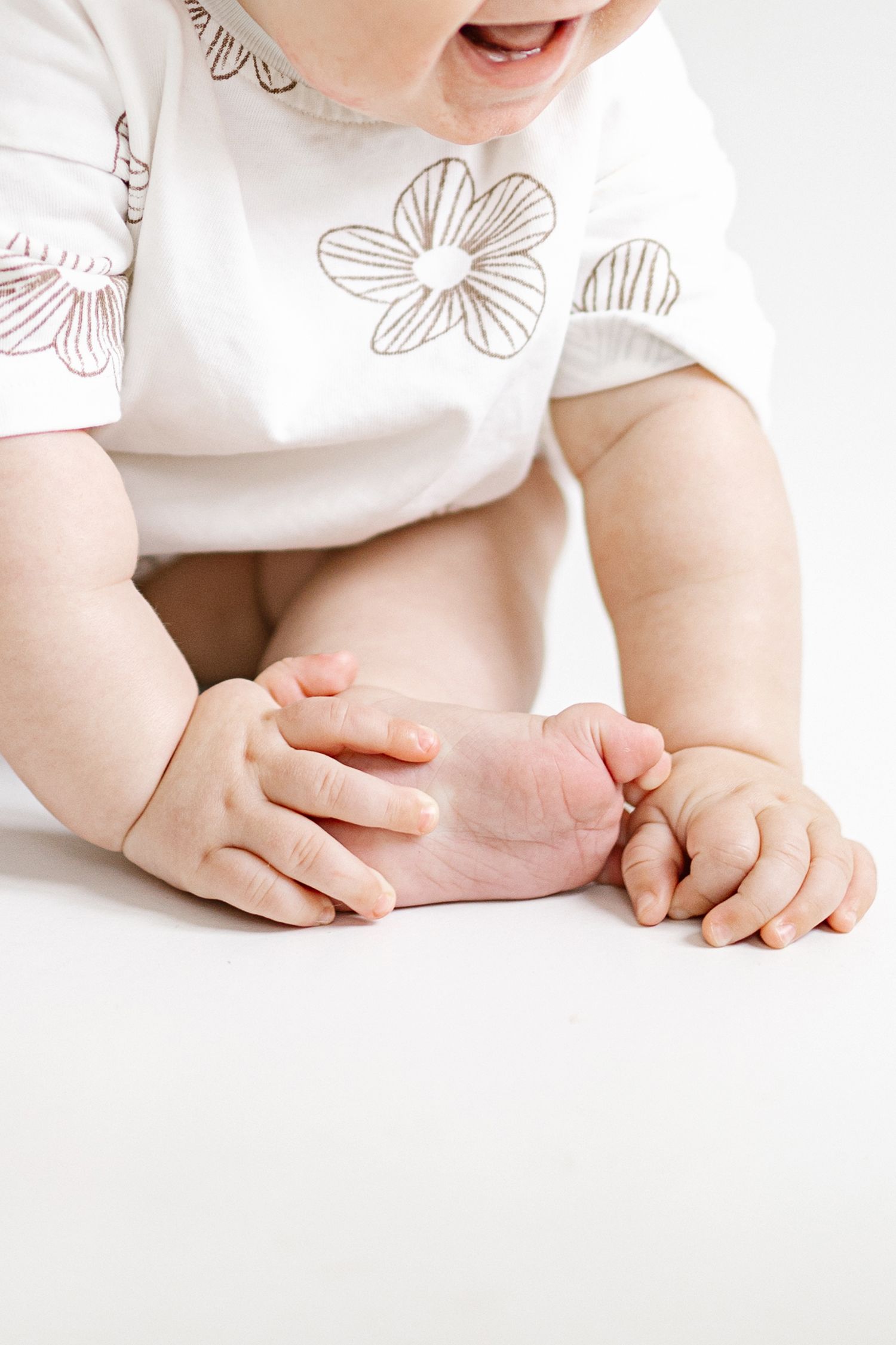 Cute baby in white floral onesie sitting and holding their foot with chubby hands on white background.