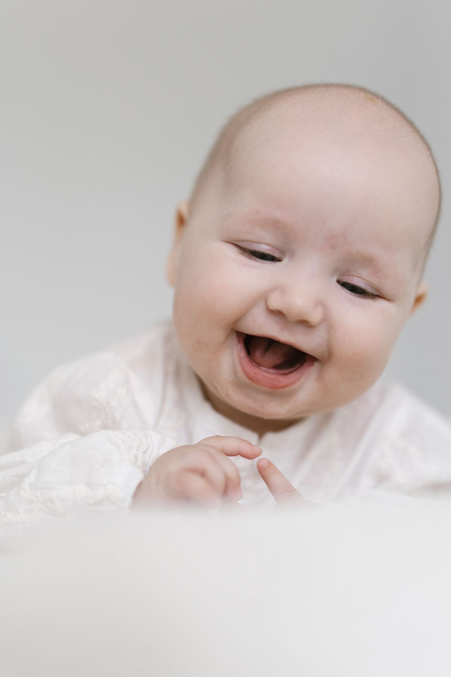 A joyful baby in a white outfit laughs with eyes closed, showing a toothless smile against a soft gray background.
