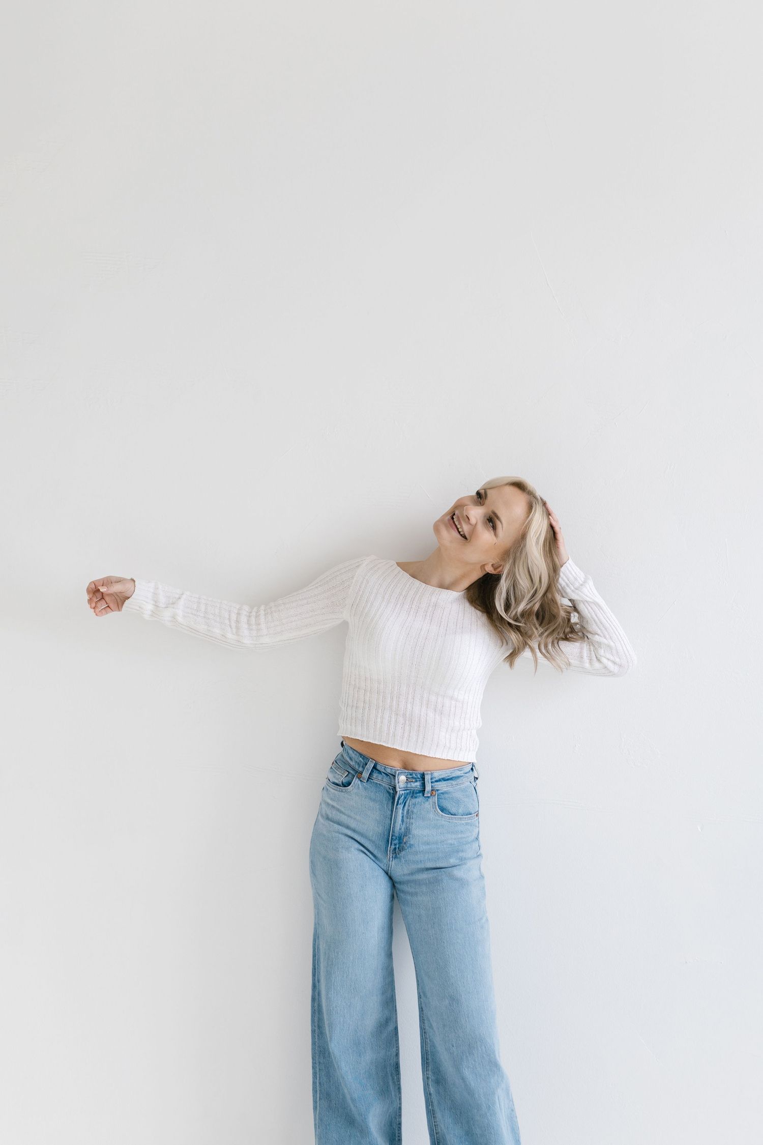Person in light sweater and blue jeans jumping joyfully against white background.