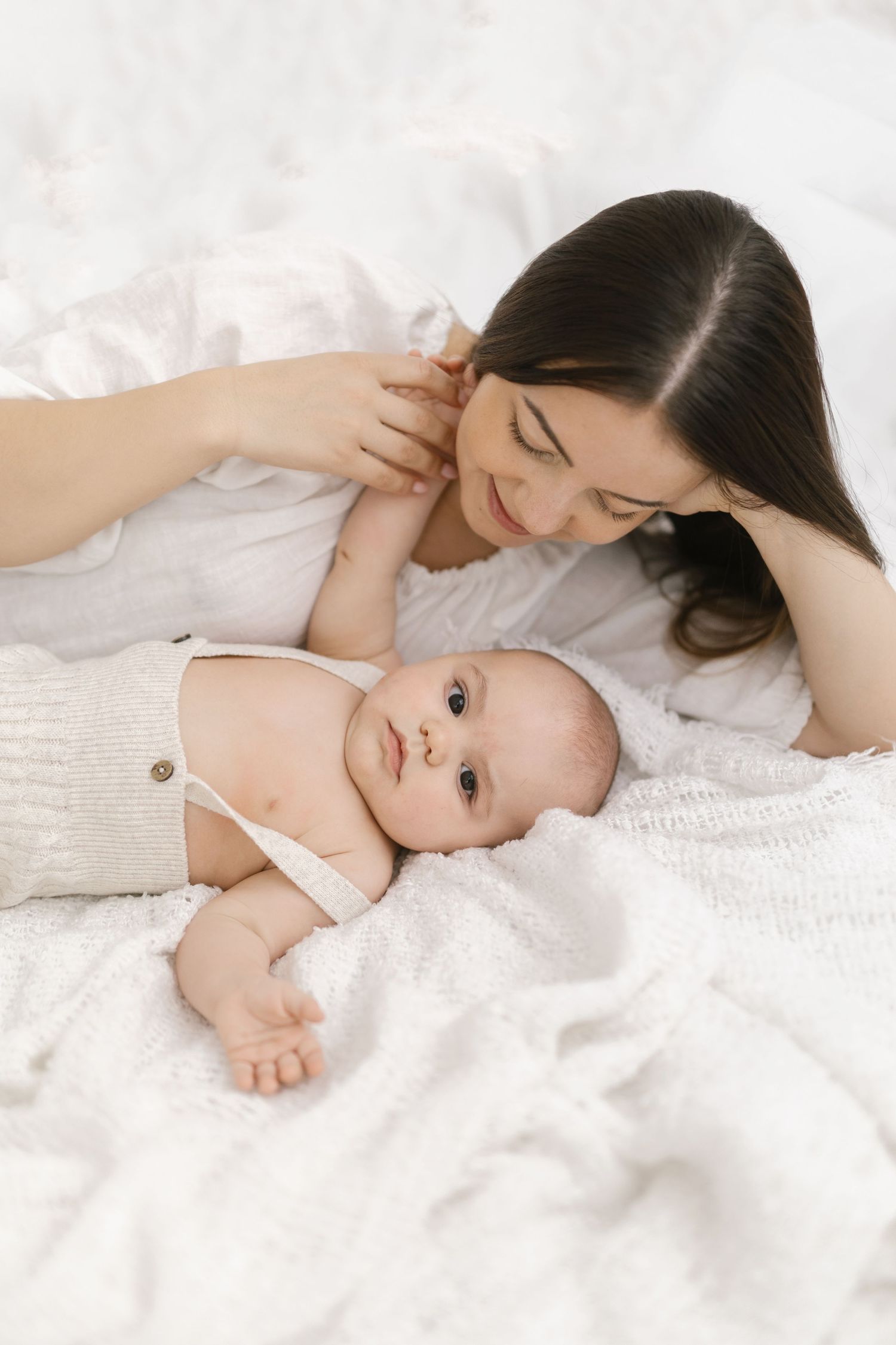 A young mother lying on a white bed gazing lovingly at her newborn baby dressed in a cream outfit.