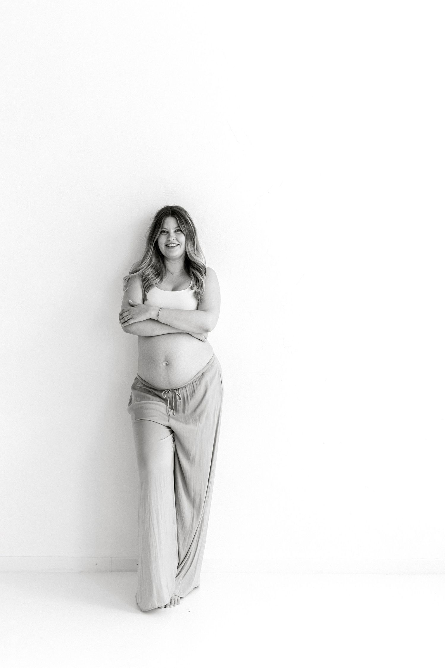 Black and white photo of a smiling pregnant woman in a crop top and wide-leg pants leaning against a white wall.
