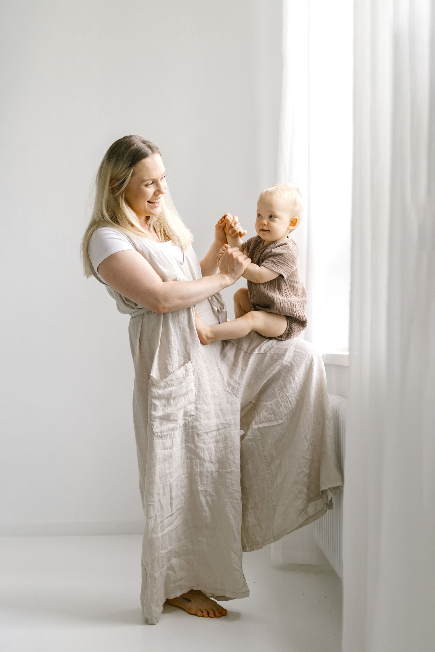 Parent and baby in light linen clothing sharing tender moments in bright white room.