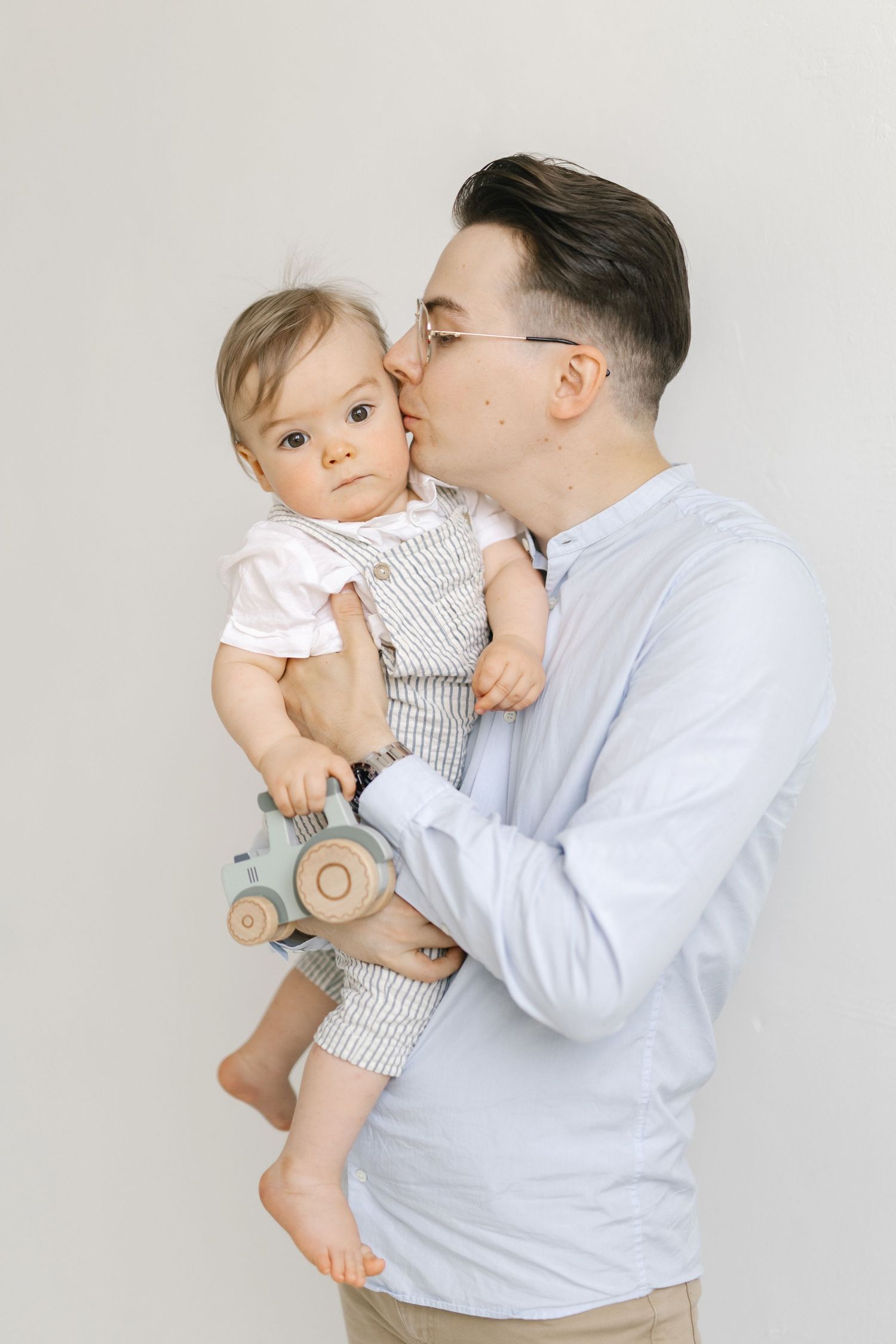 Adult giving baby in striped overalls a kiss against white background.