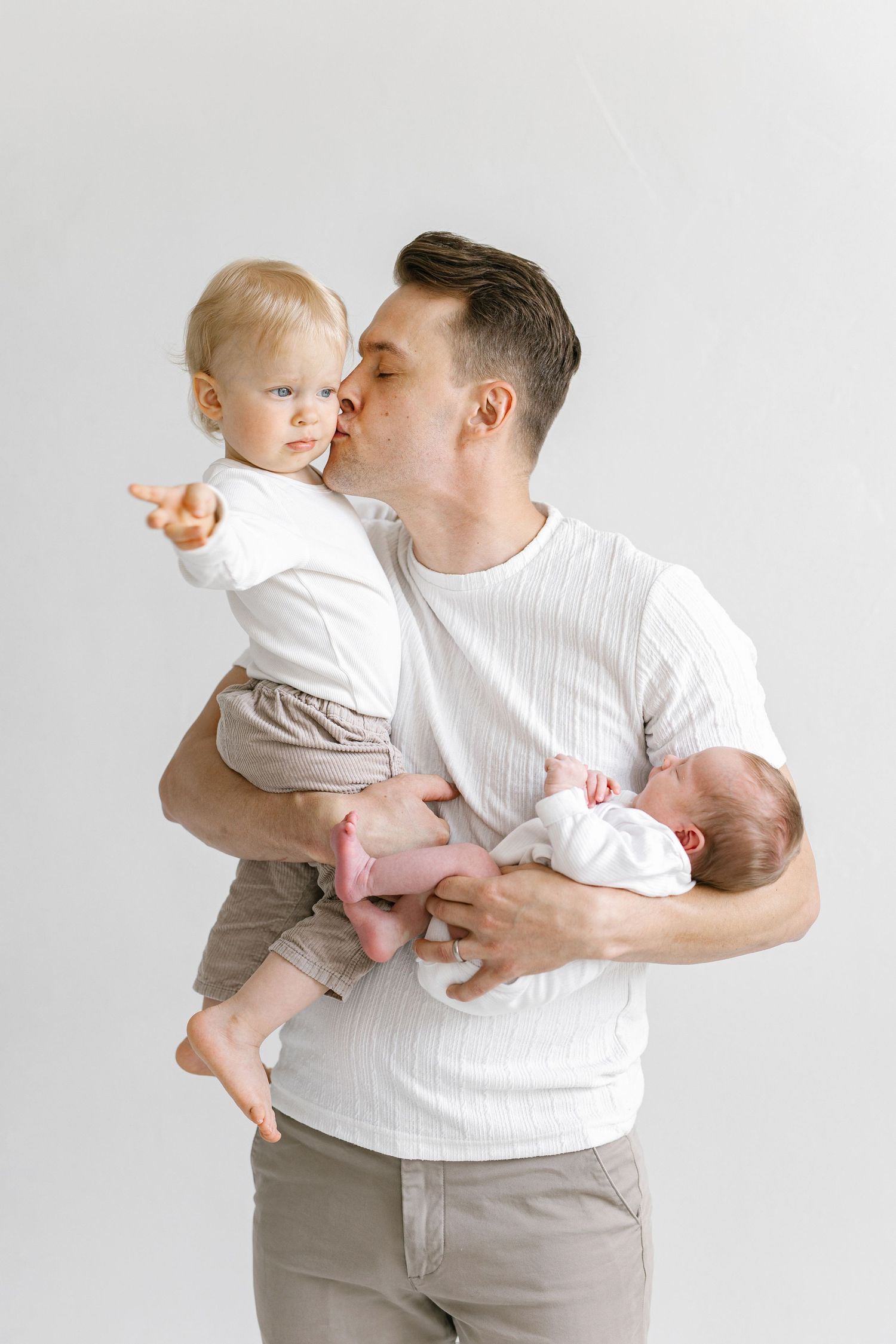 Father holding toddler and newborn baby, kissing older child on cheek, studio white background.