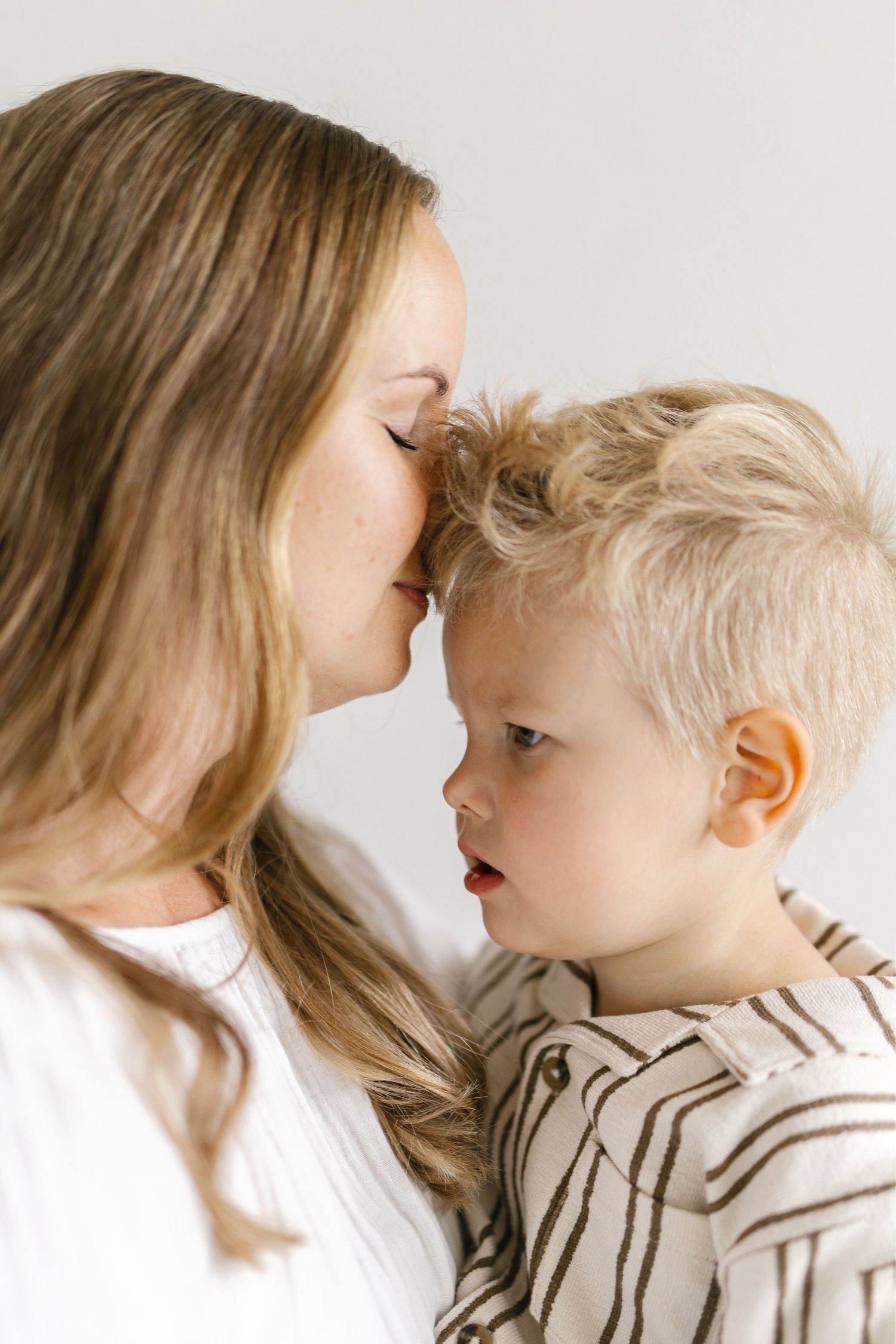 A woman with long blonde hair gently presses her forehead to a toddler boy in a striped linen shirt in a tender, bright indoor portrait.