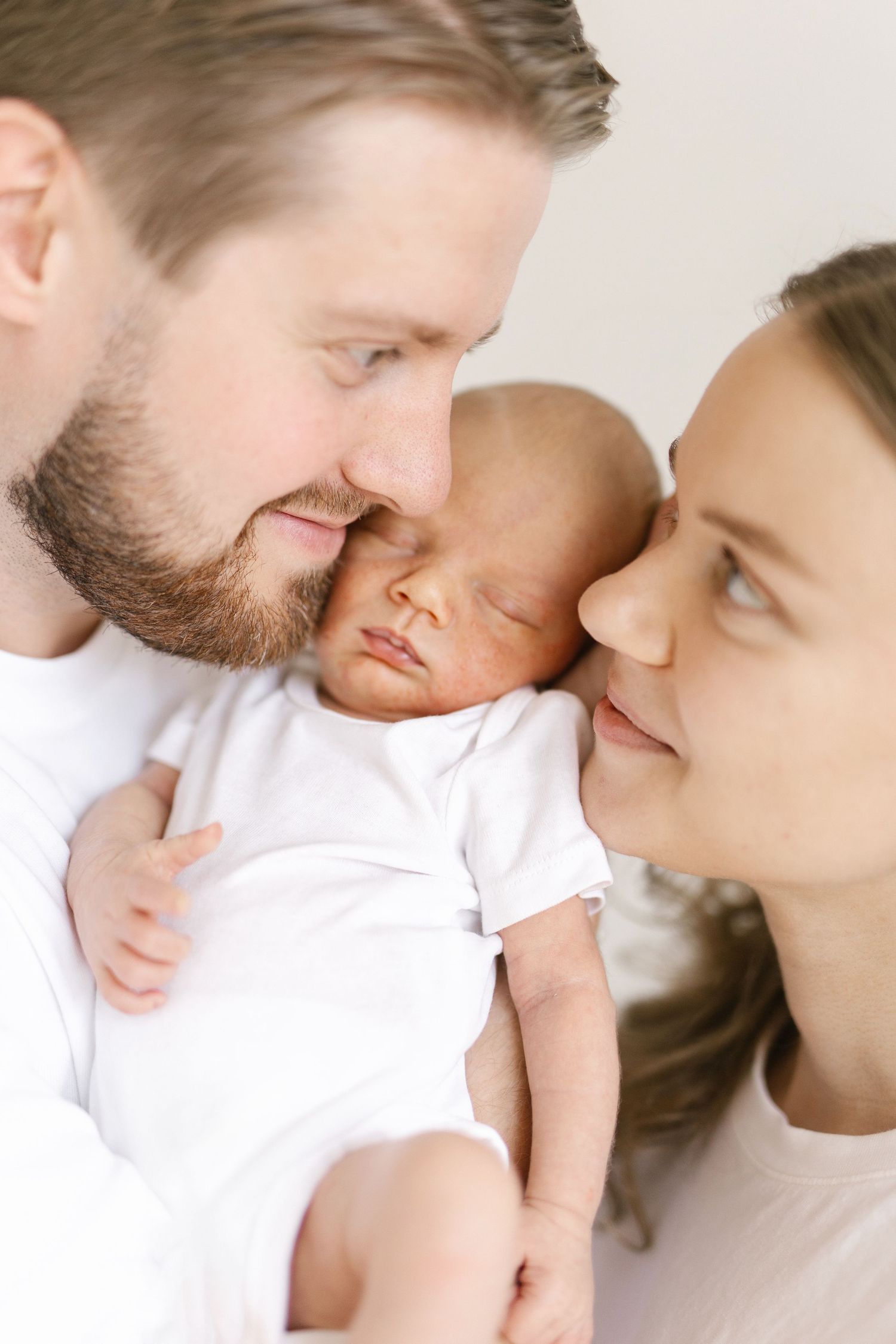 New parents lovingly hold their sleeping newborn baby dressed in white, sharing a tender family moment.