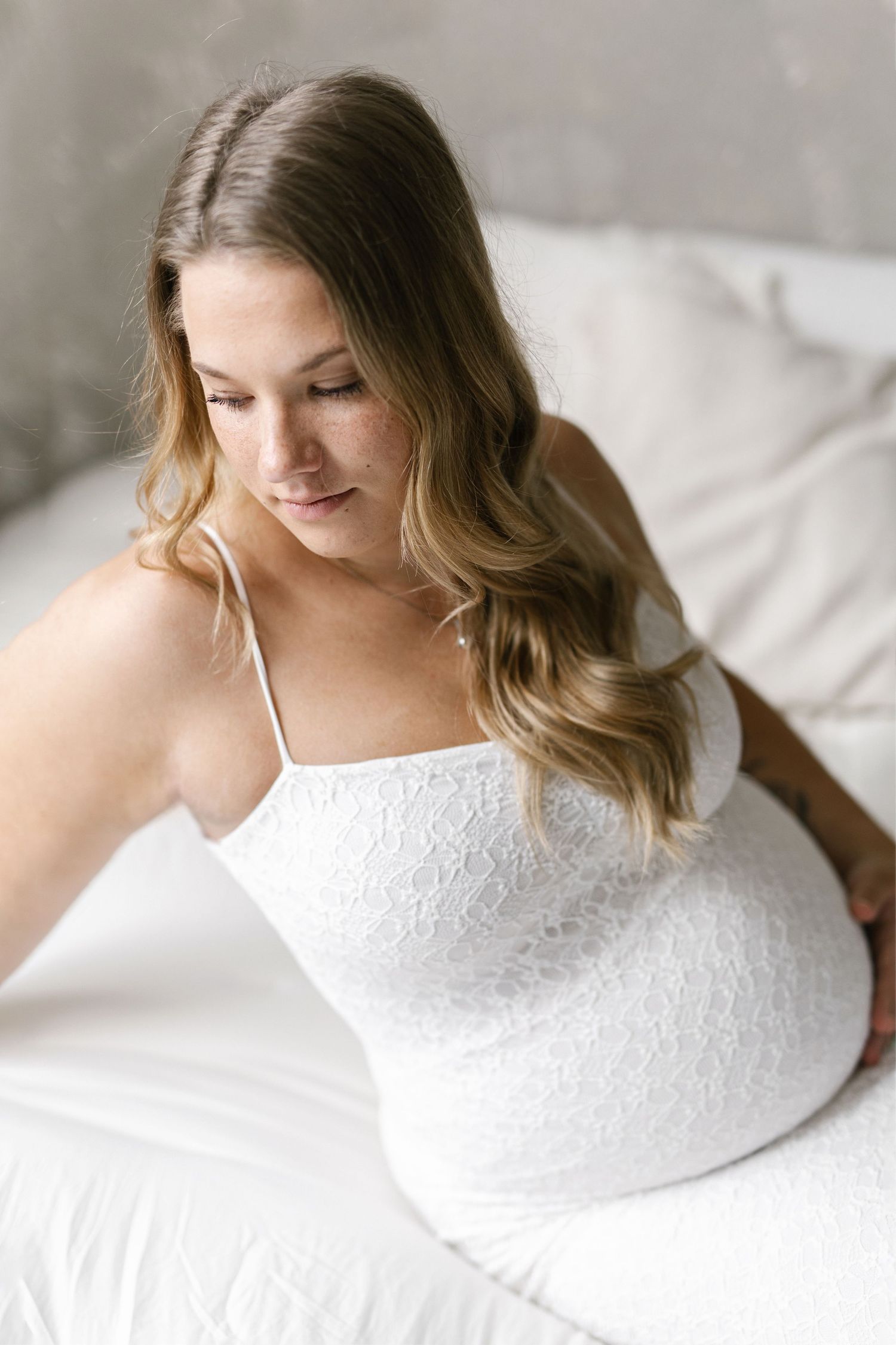 Pregnant woman in white lace dress sitting on bed, looking down at baby bump during maternity photo session.