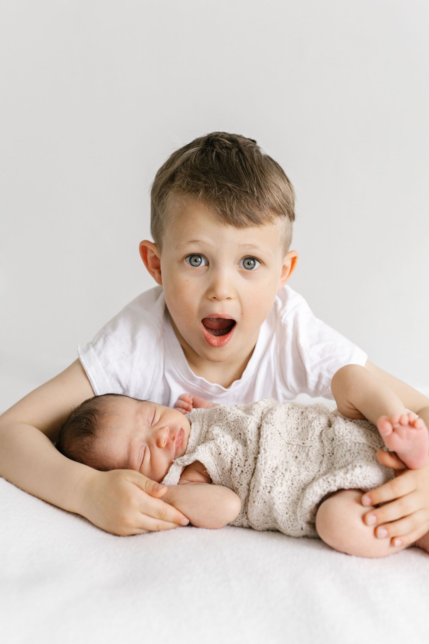 Excited young boy with blue eyes holds sleeping newborn baby sibling, white background, family portrait.