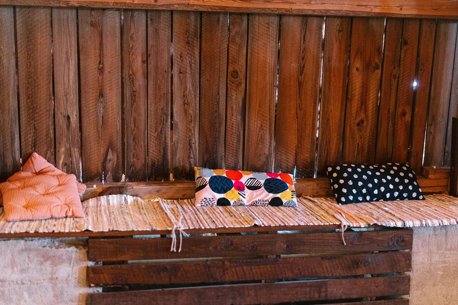 Rustic wooden bench with colorful decorative pillows on a striped mat against a wood plank wall.
