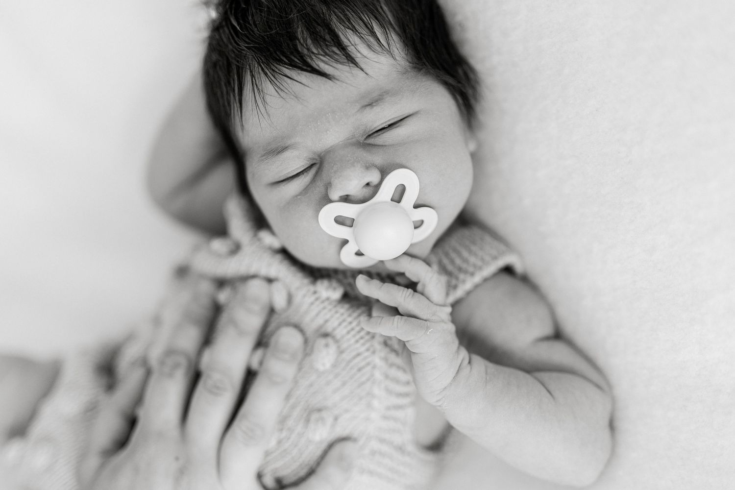 Black and white photo of a sleeping newborn baby holding a pacifier, wrapped in a knit blanket.