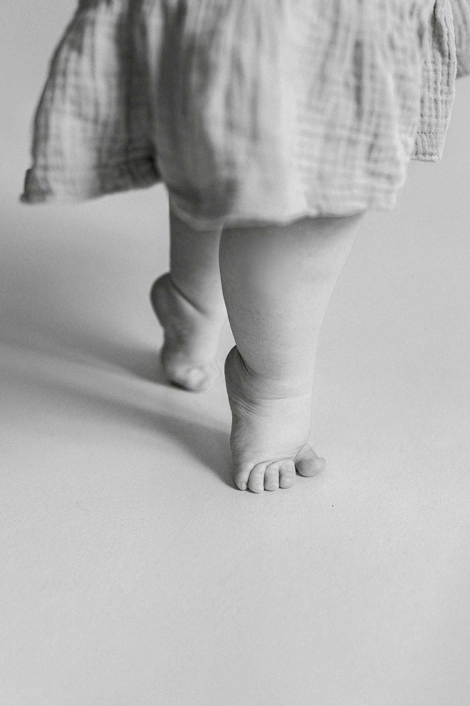Black and white photo of a baby's chubby feet taking first steps, wearing a soft muslin dress.