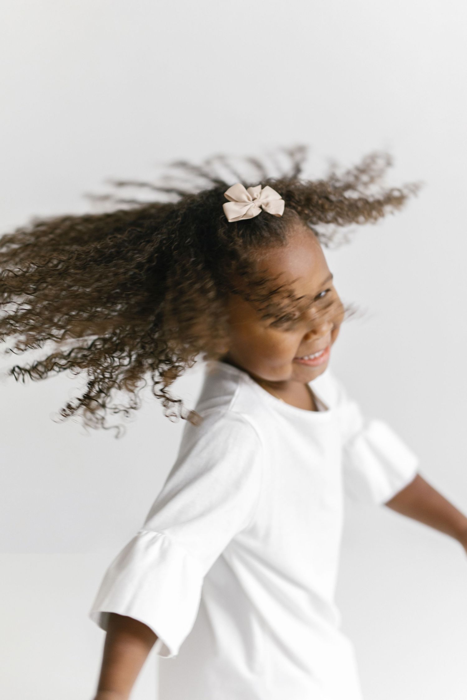 A joyful young girl in a white dress twirls, her curly ponytail flying, adorned with a pink bow hair clip.
