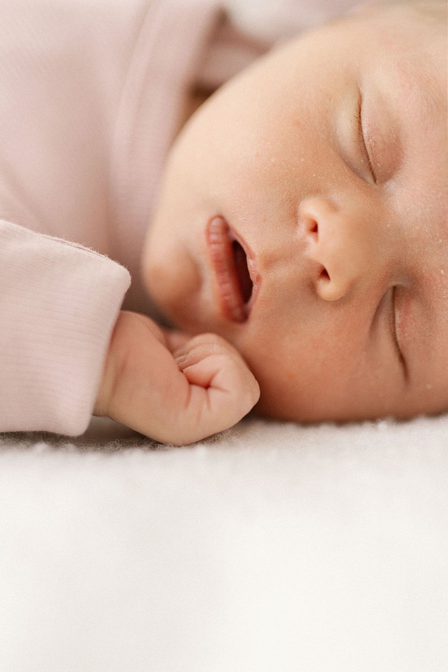 A peaceful newborn baby sleeping soundly on a white fluffy blanket, wearing a soft pink outfit with tiny clenched fists.