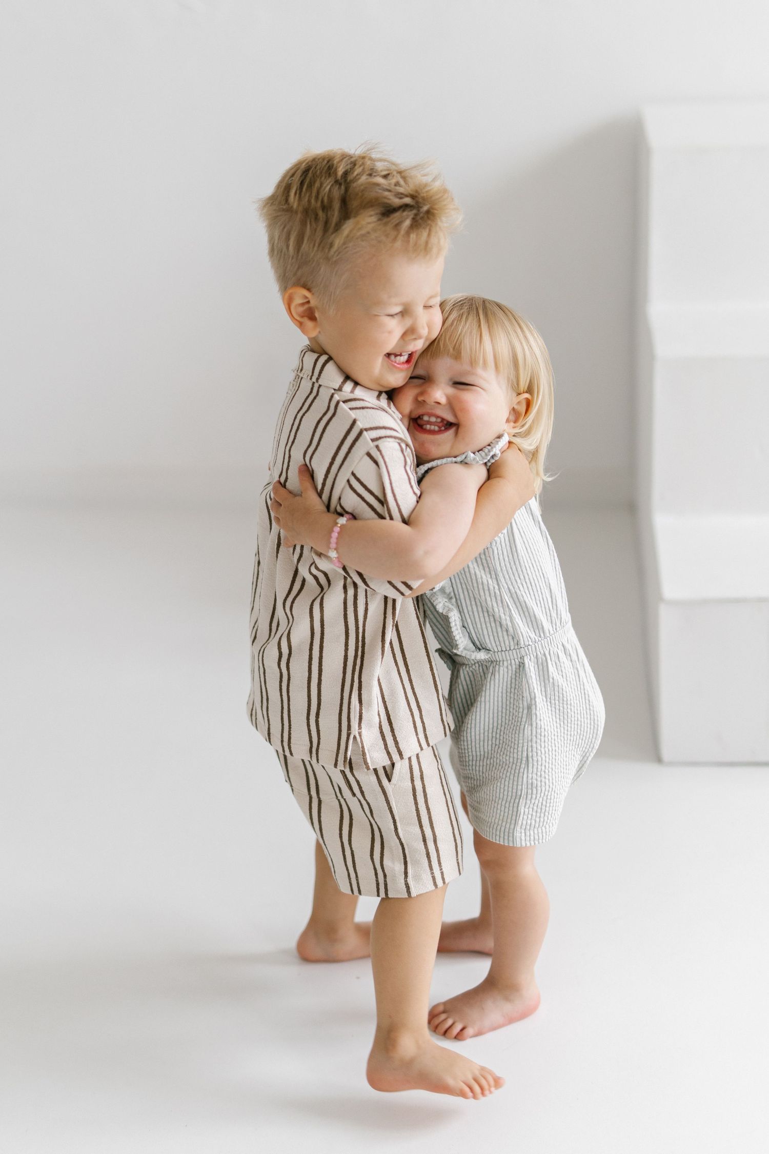 Two toddlers hugging and laughing in striped linen outfits against a white background.